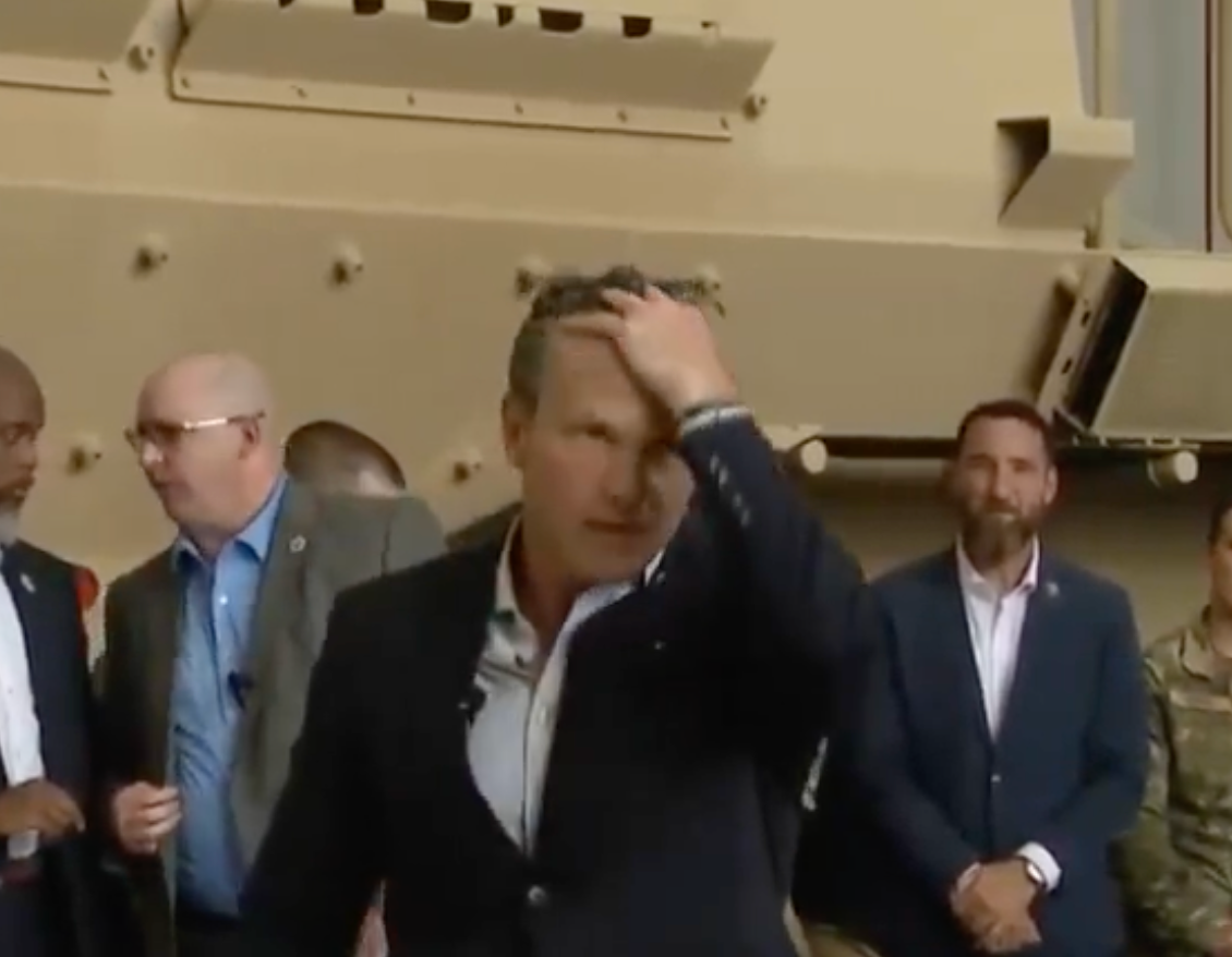 En mann i dress tar på håret hans, omgitt av folk i forretnings- og militærantrekk. De ser ut til å være i en organisert setting