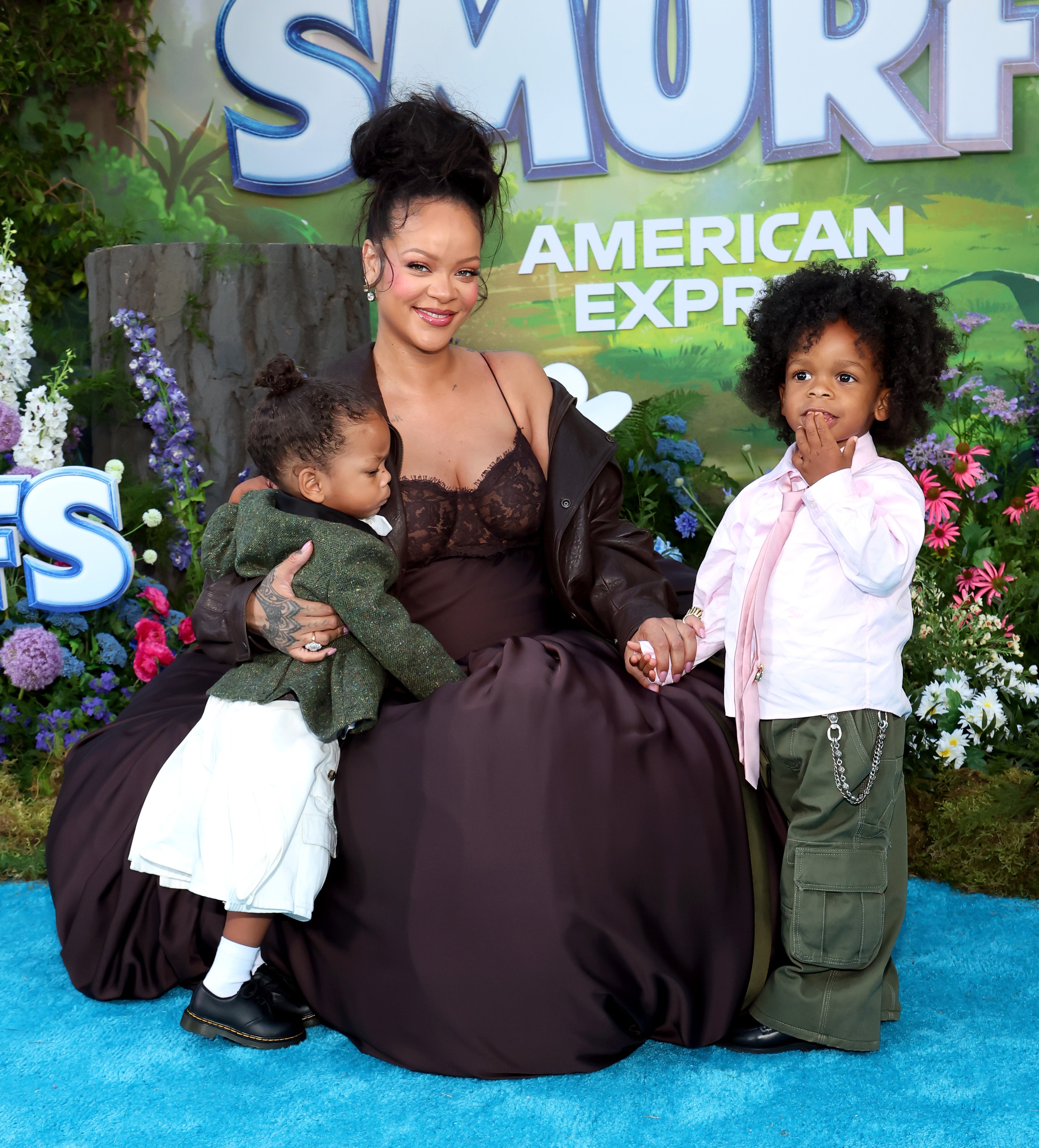 A person in a stylish dress sits on a blue carpet with two children at an event. The backdrop features the word "Smurfs."