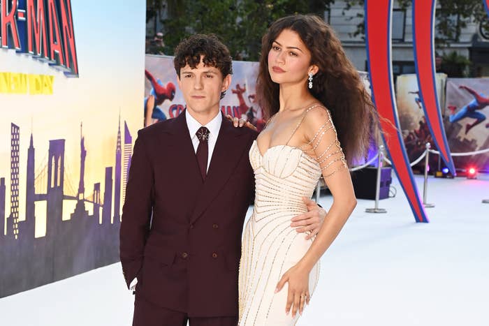 Two individuals on a red carpet; one in a burgundy suit, the other in a white strapless gown with chain details
