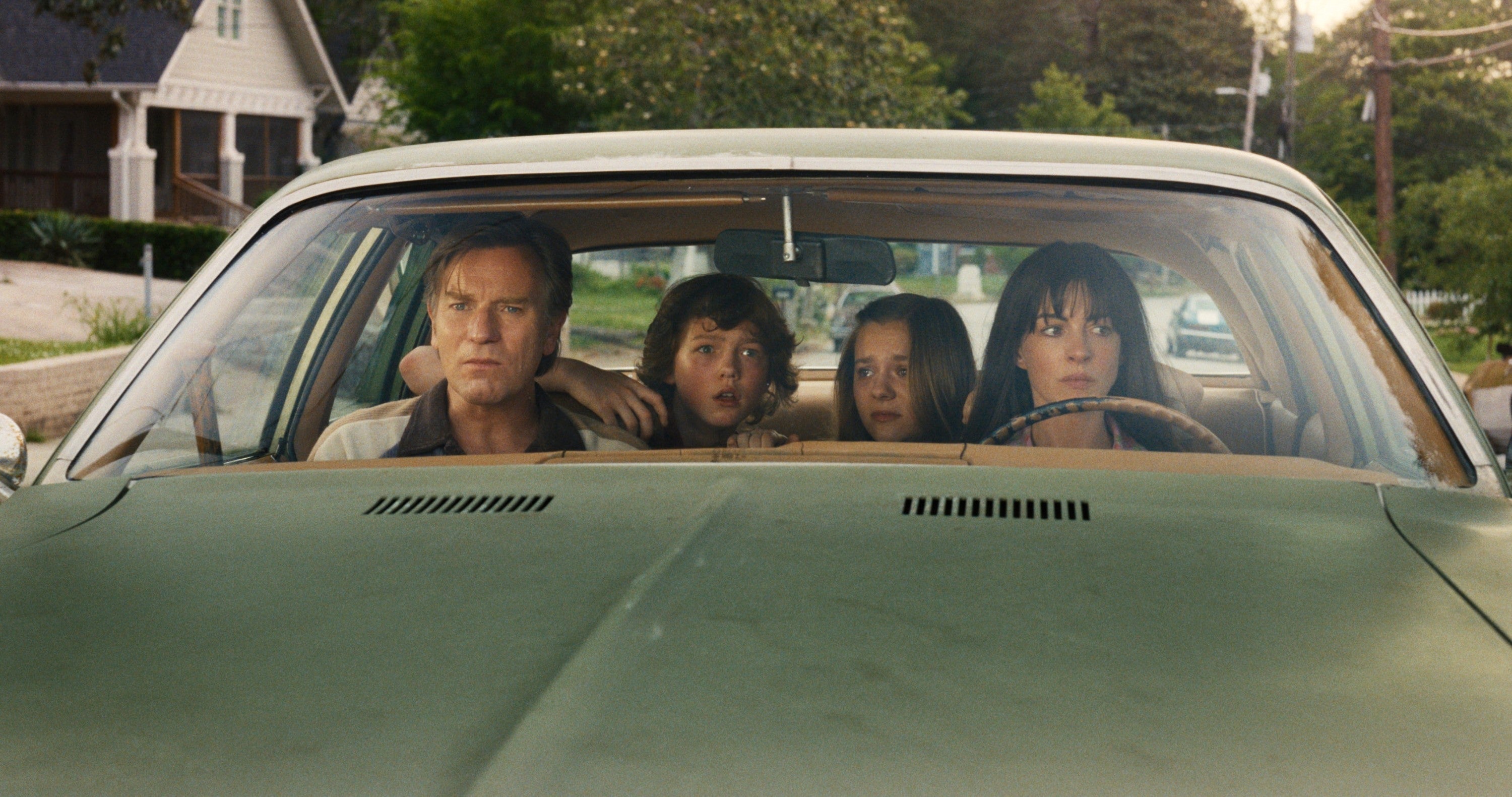 Four people sitting tensely in an old car, staring out the windshield, seemingly concerned or focused