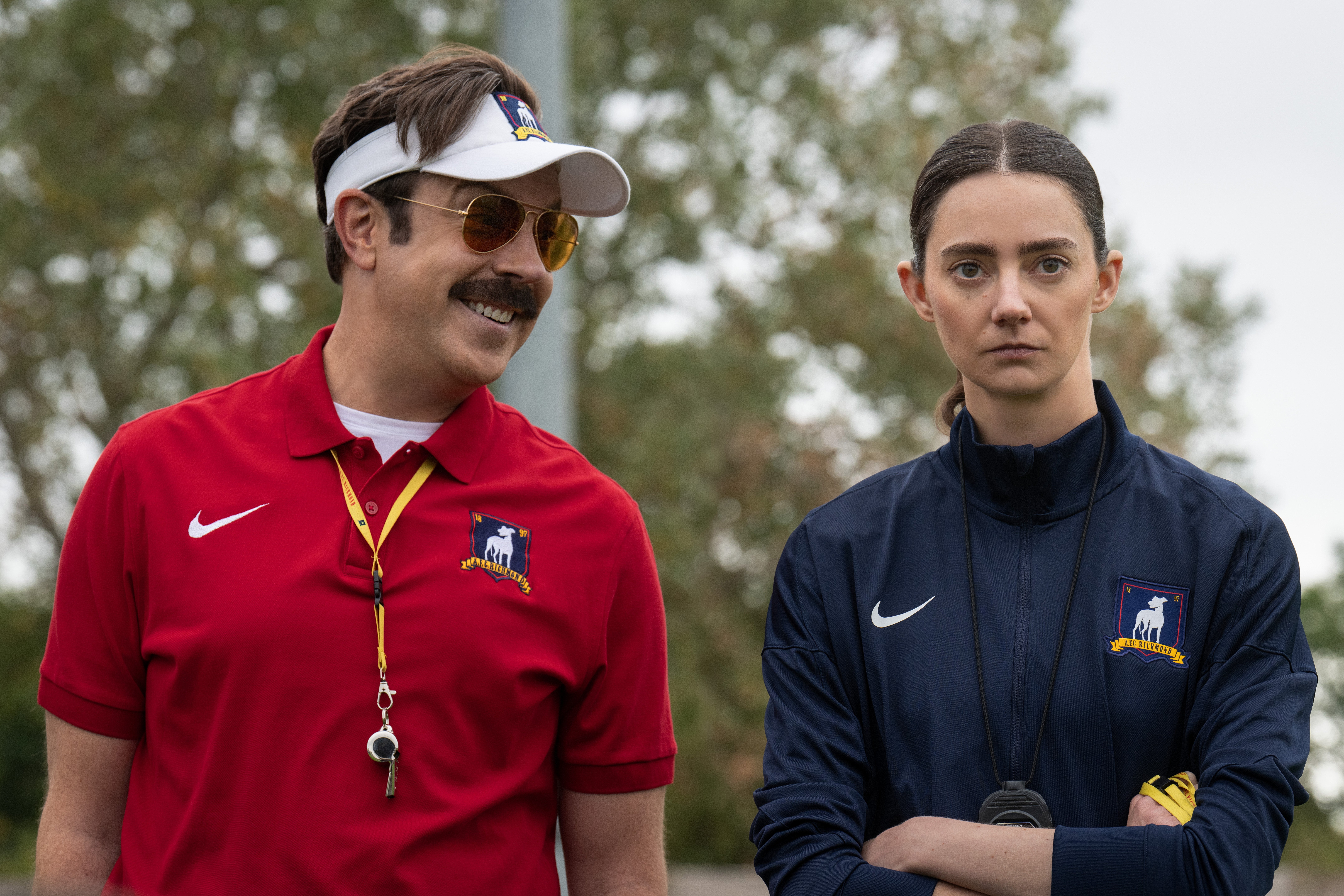 Two actors on a set. The man in a sports visor and red polo smiles, while the woman in a blue tracksuit looks serious
