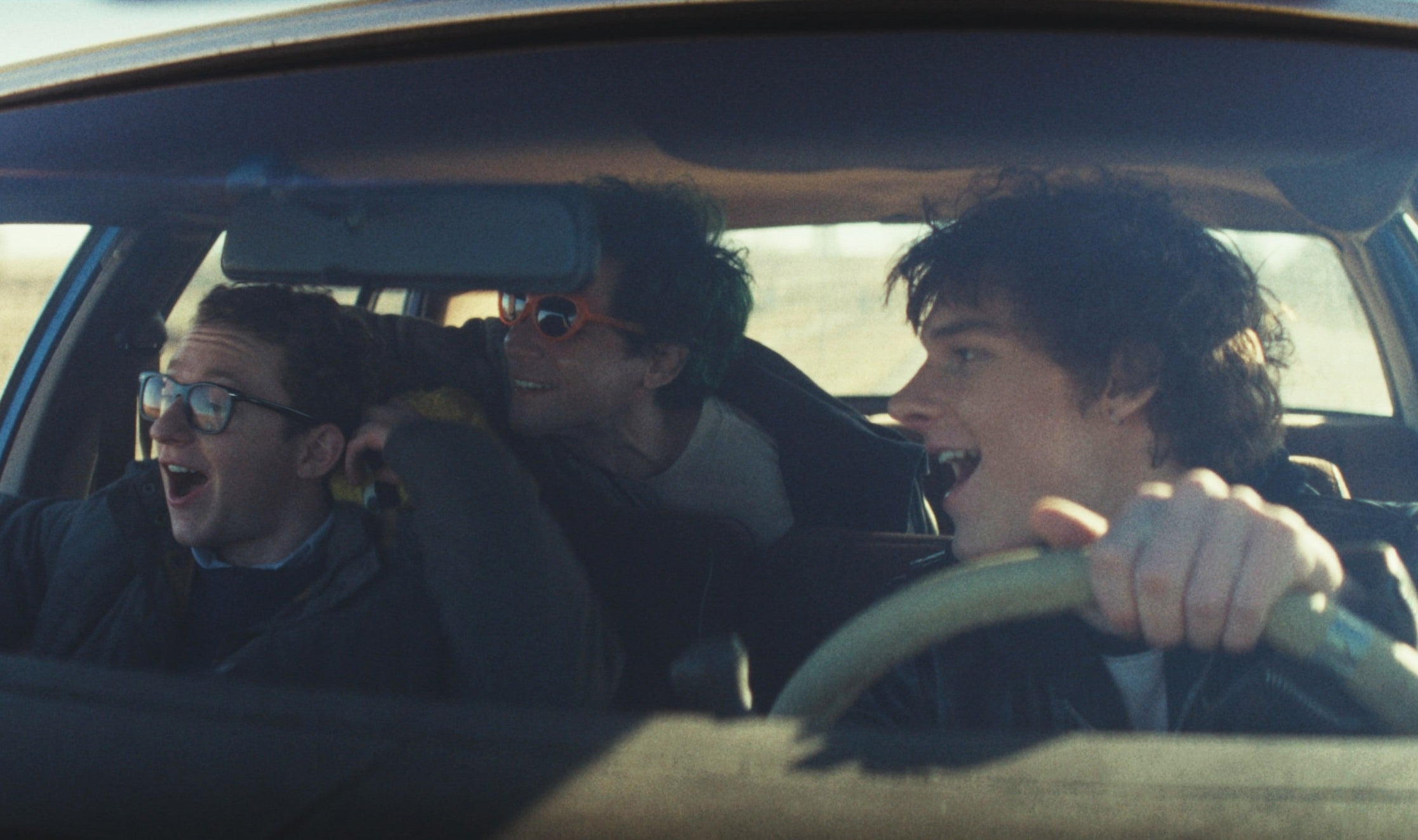 Three people are excitedly singing in a moving car, with one person driving and two passengers joining in