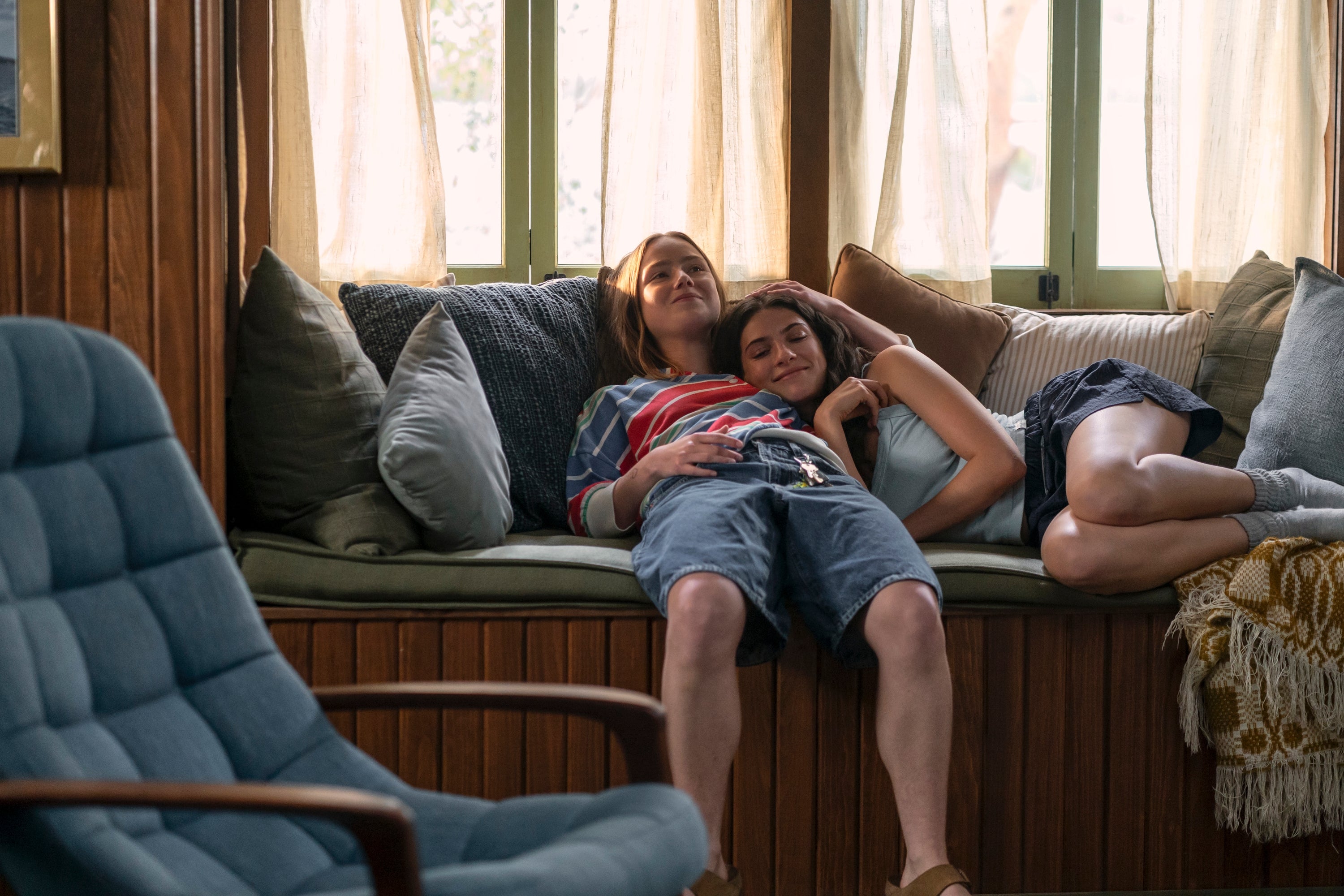 Two people relax on a cushioned window seat. One leans on the other's shoulder, both appear content and cozy