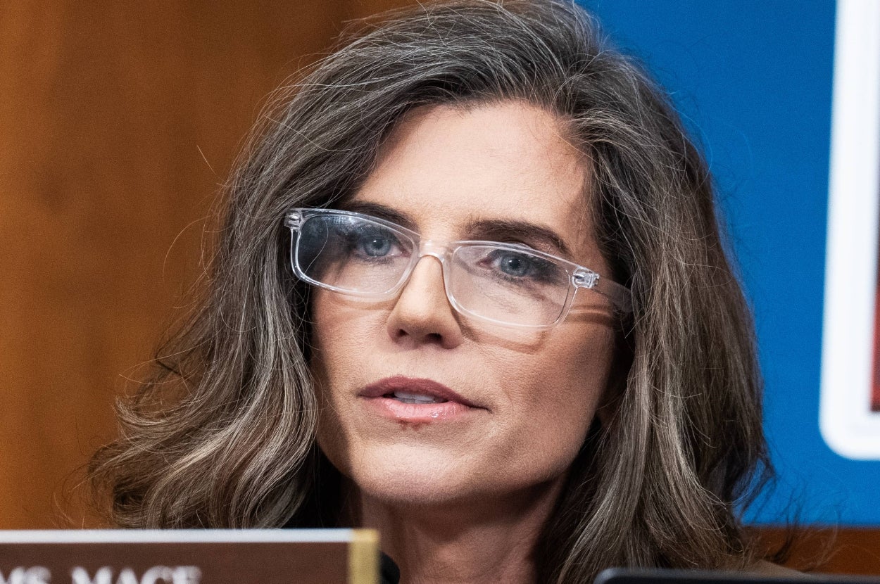 Person with wavy hair and glasses speaks at a formal event, seated behind a microphone with a focused expression