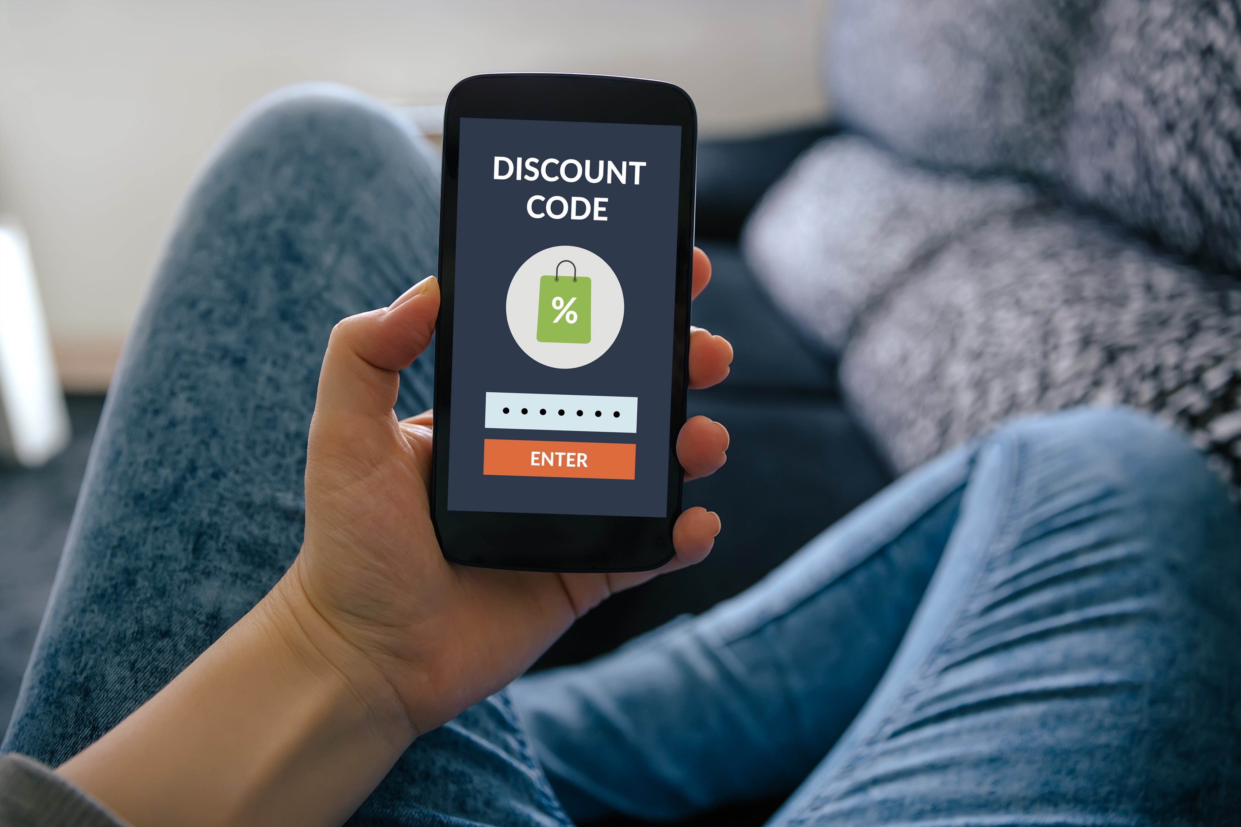 Person holds a smartphone displaying a "Discount Code" surface with a percent awesome and an "Enter" button. Jeans and sofa disposable successful the background