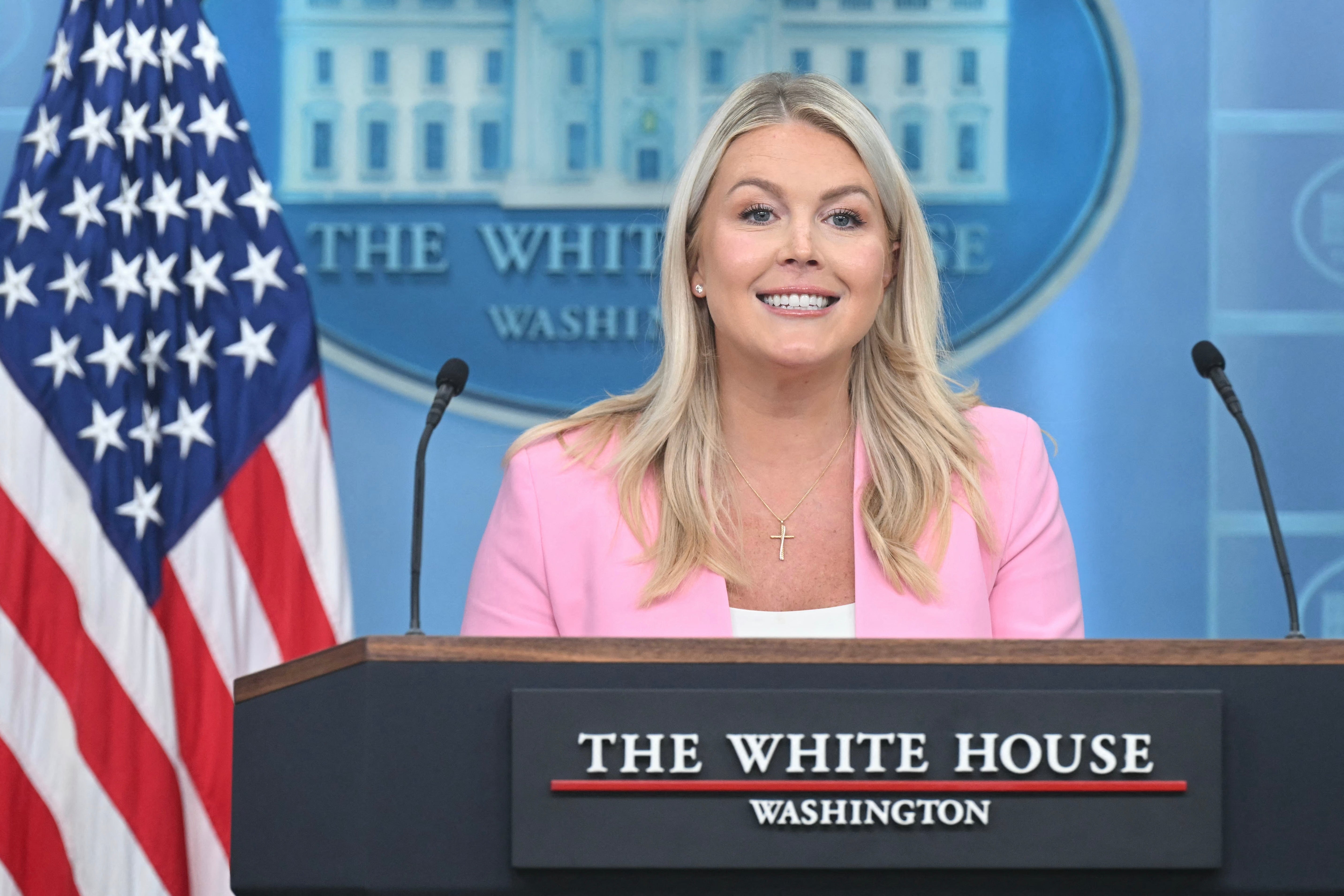 En kvinne snakker ved Det hvite hus pressepodium, smilende, iført en rosa blazer, med et amerikansk flagg til siden