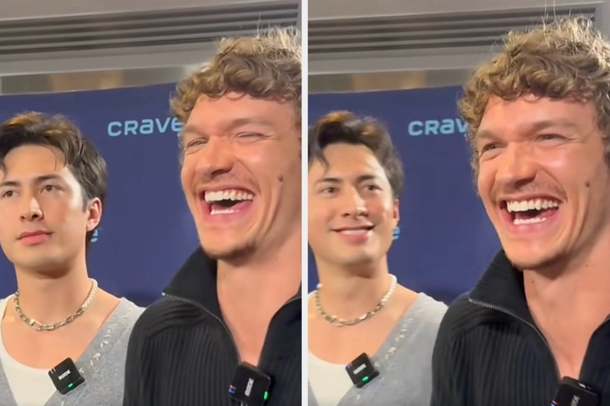 Two people laughing during an interview; one wears a black jacket, the other a gray shirt with a necklace