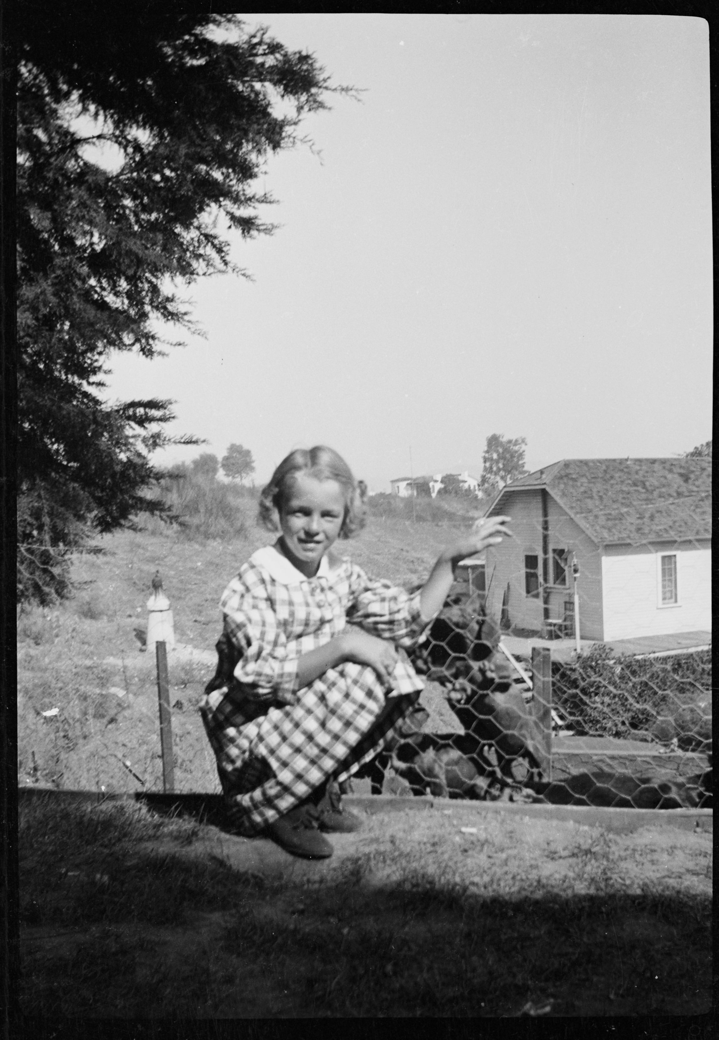 Marilyn Monroe as a child crouching by a wire fence, wearing a checked dress, with houses and trees in the background