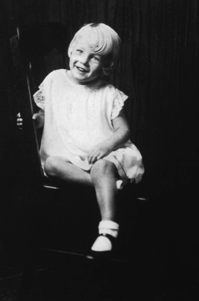 Marilyn Monroe as a child with short hair sits on a chair, wearing a light dress and socks with sandals