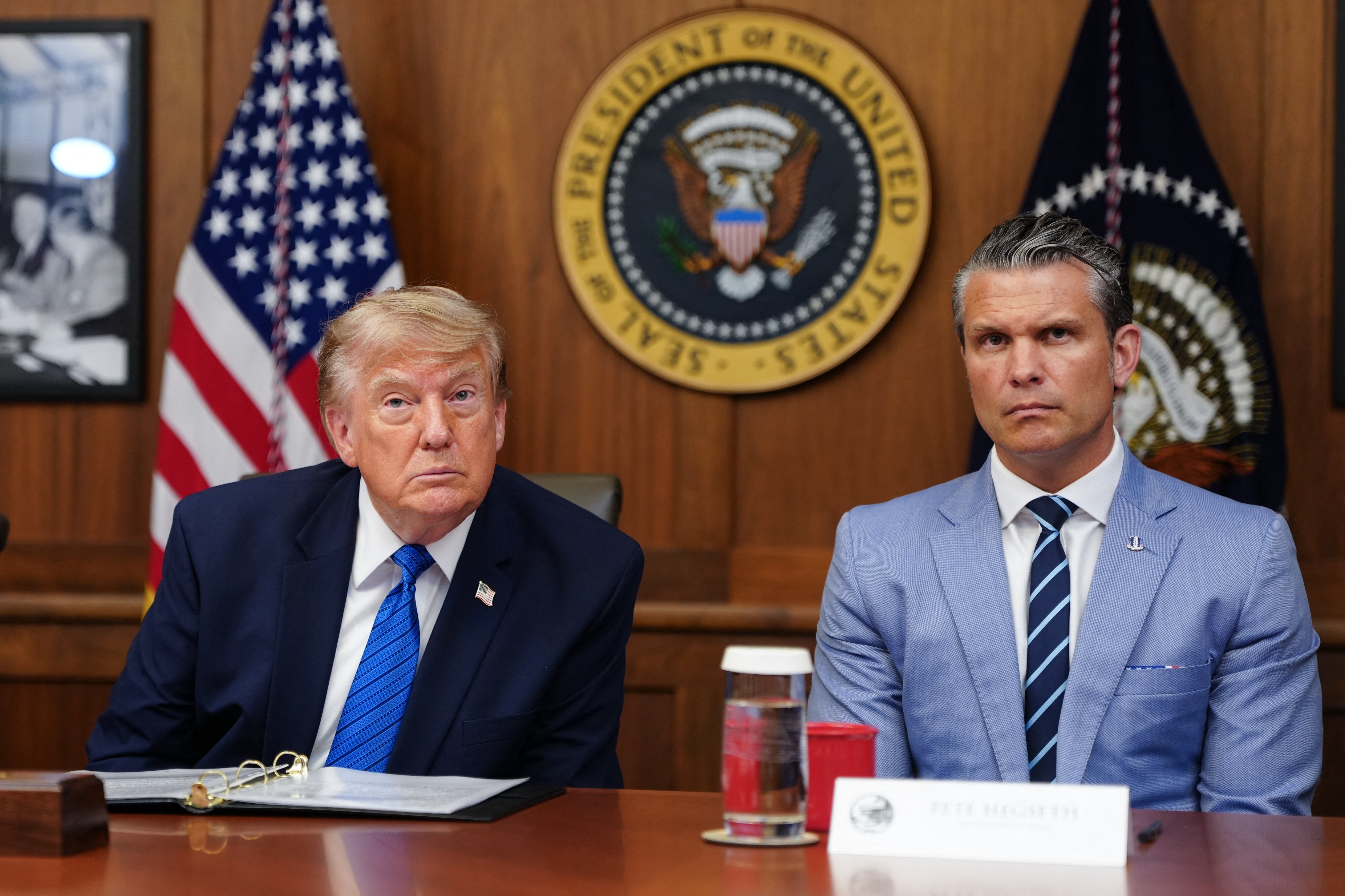 To menn sitter ved et bord i formelle omgivelser med det amerikanske presidentstempelet i bakgrunnen. En mann har på seg dress og slips; den andre en lettere dress