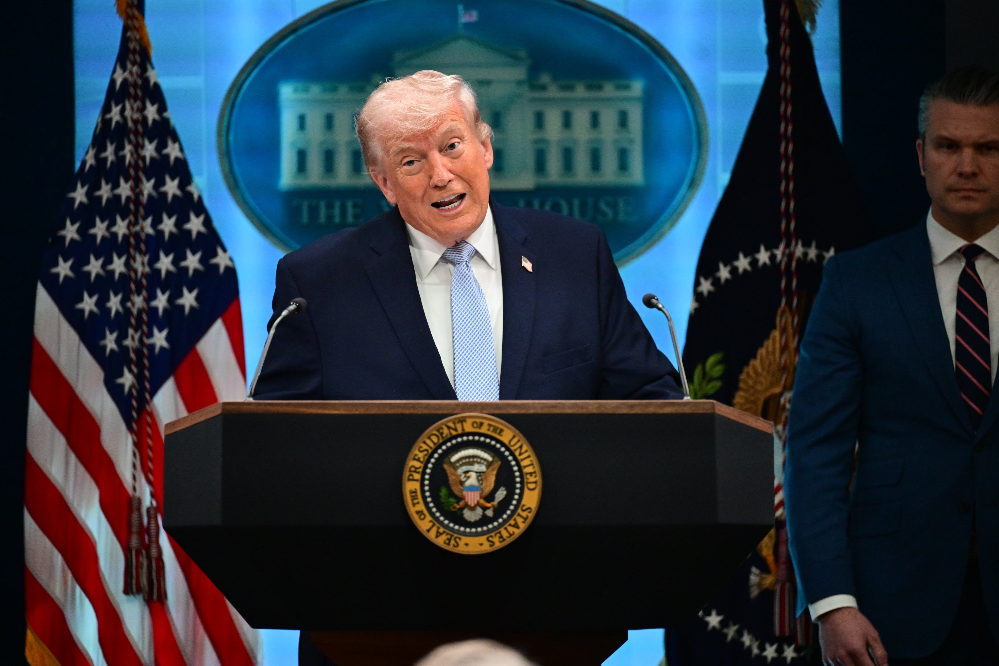 En person snakker på et podium med et presidentstempel, flankert av amerikanske flagg og et offisielt bakteppe, med en annen person som står i nærheten