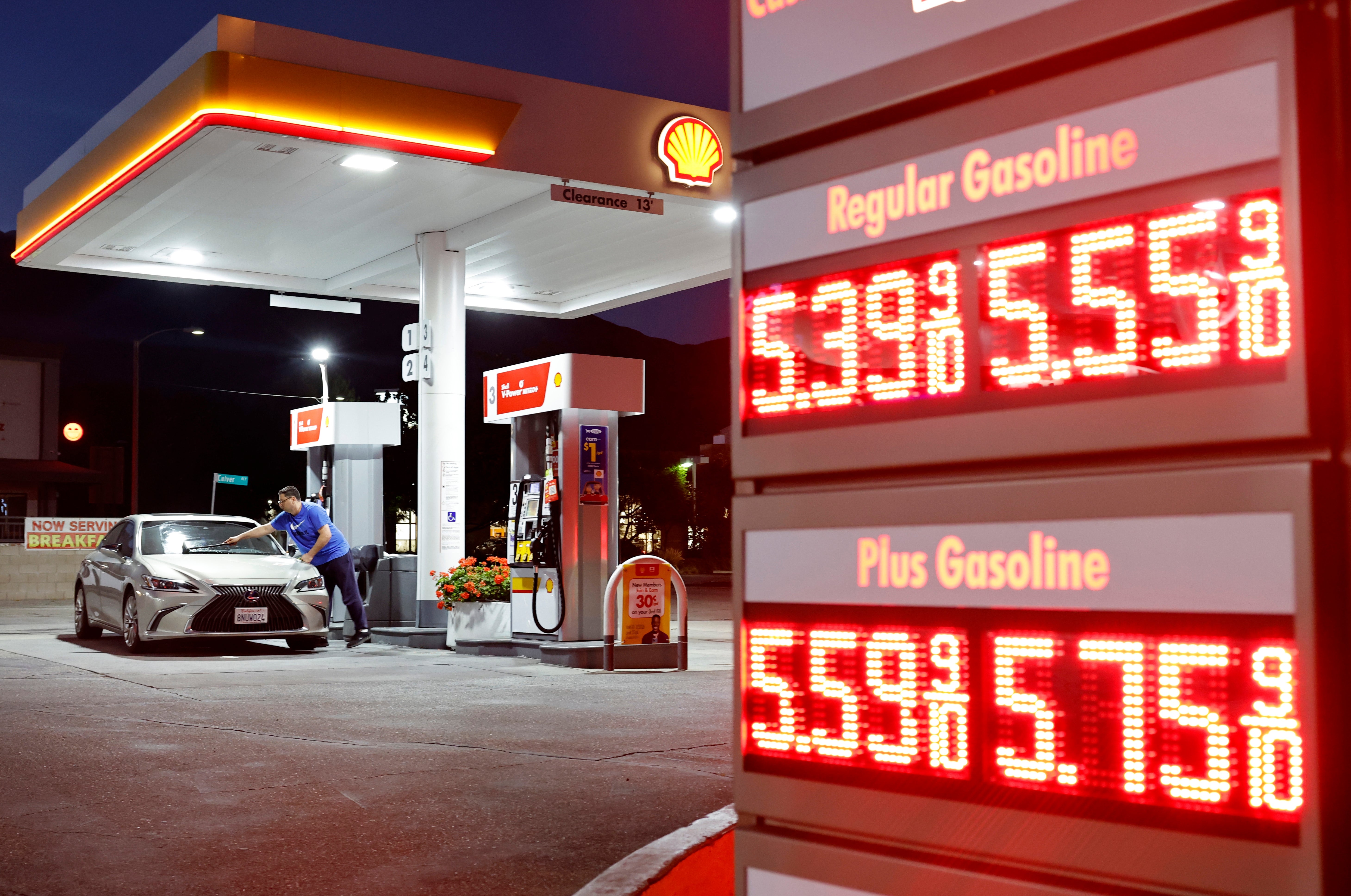 Person som fyller drivstoff på en bil på en bensinstasjon om natten; prisene som vises for vanlig og pluss bensin overstiger $5,50 per gallon