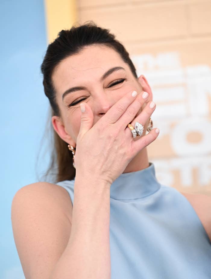 Person with long hair laughs, covering their mouth with a hand adorned with rings and polished nails, wearing a high-neck sleeveless top