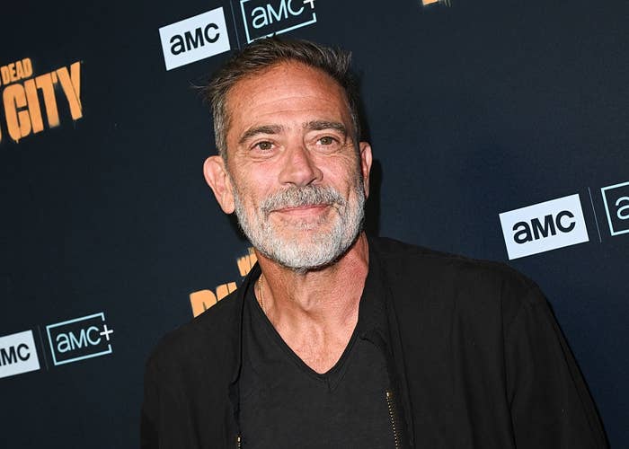 Man with short hair and beard on a press event red carpet wearing a black jacket and shirt, standing in front of AMC-themed backdrop