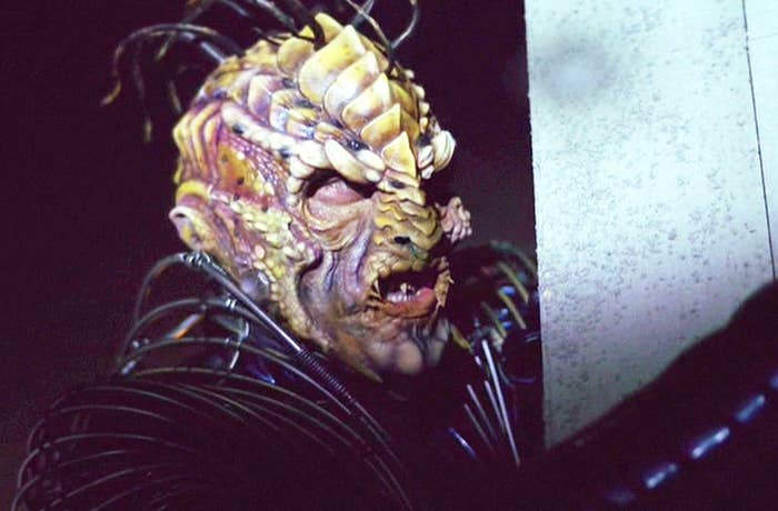 A person in elaborate alien makeup with a textured, horned mask and coiled wire costume, peering intensely from behind a corner