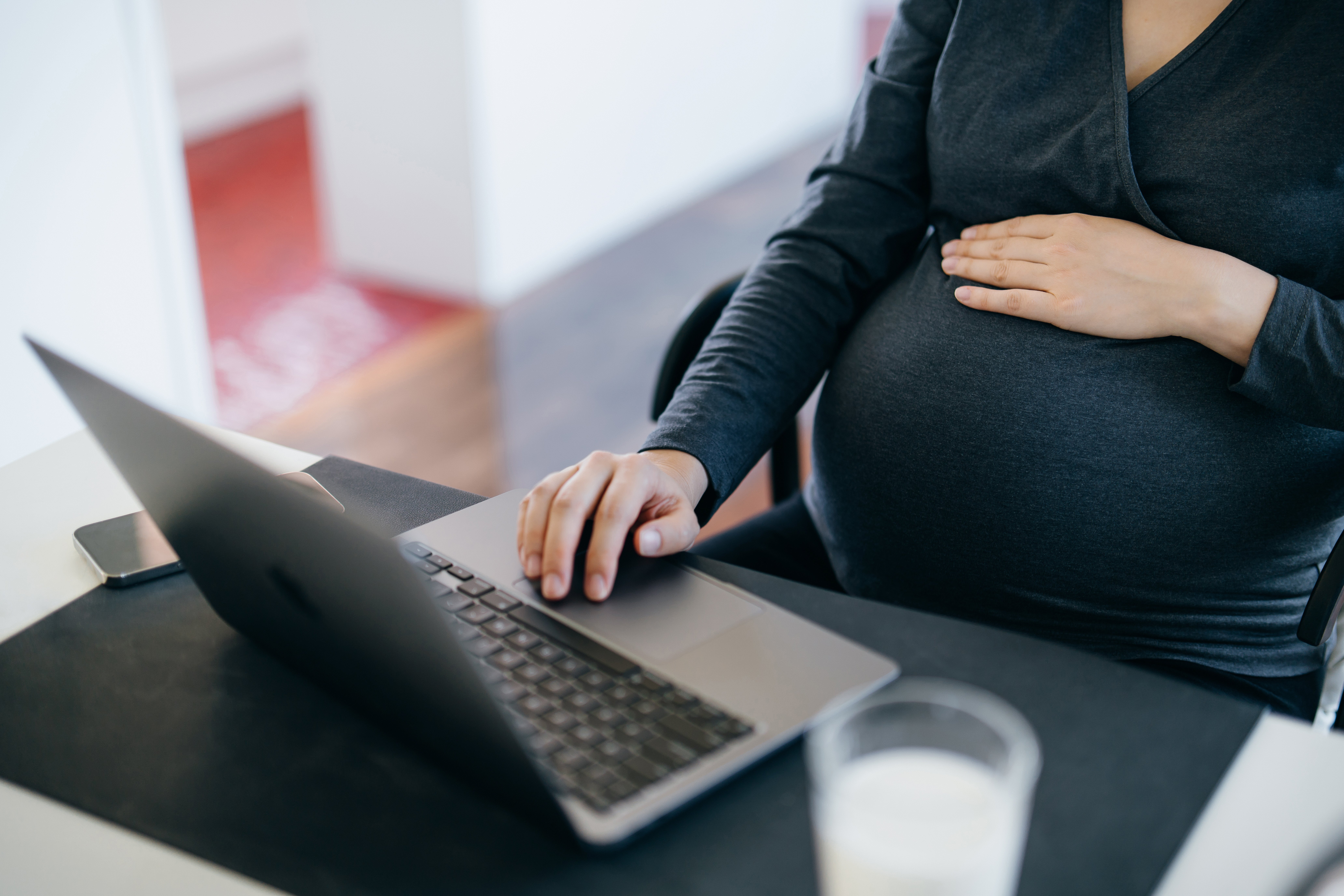 Pregnant person sitting at a desk, one hand on their belly and the other on a laptop, with a glass of milk nearby