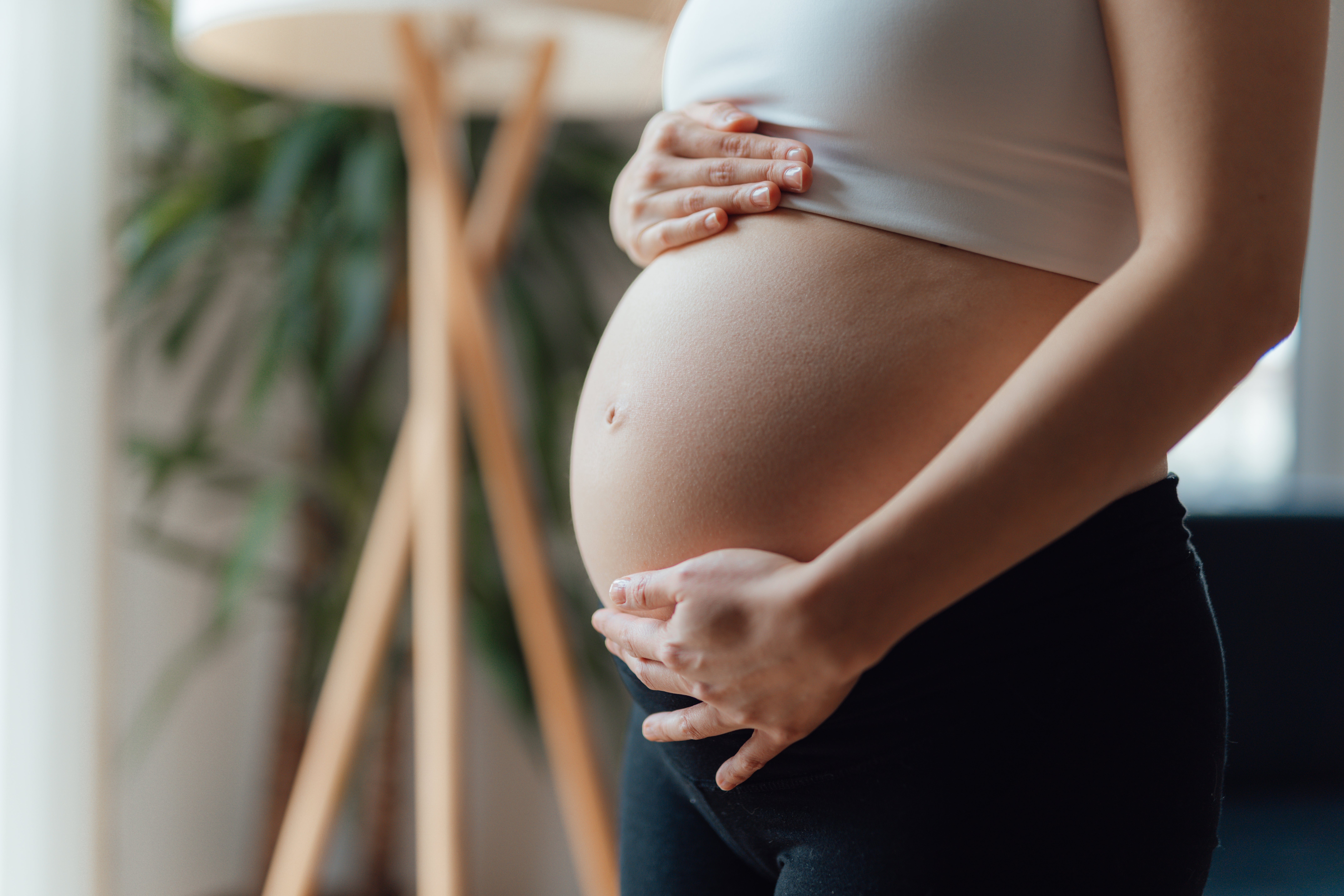 A pregnant person in a sleeveless top gently cradles their belly, standing indoors with a plant in the background