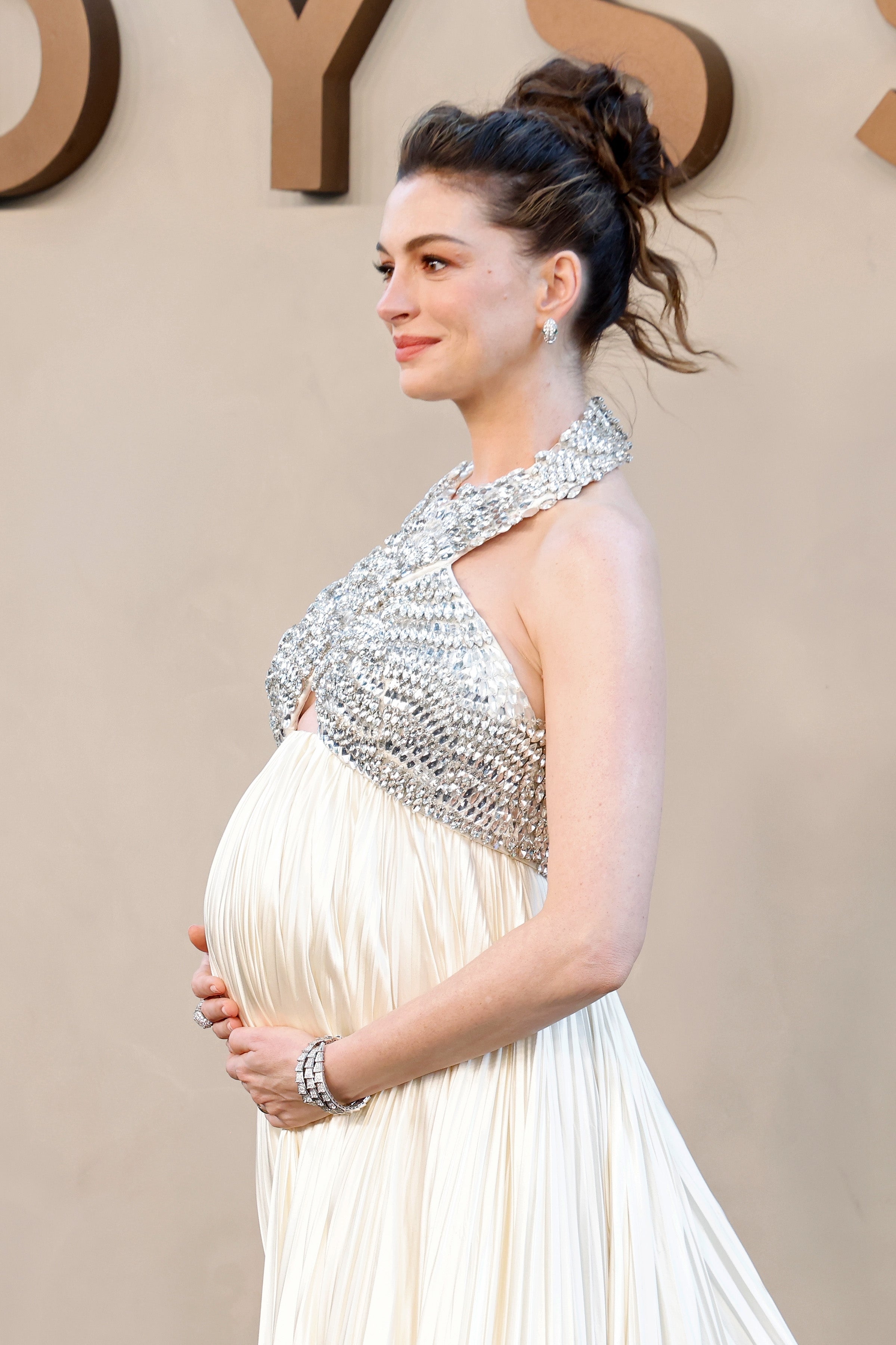 Person in elegant, flowing gown with an embellished halter top, cradling pregnancy bump at a celebrity event. Hair styled in an updo