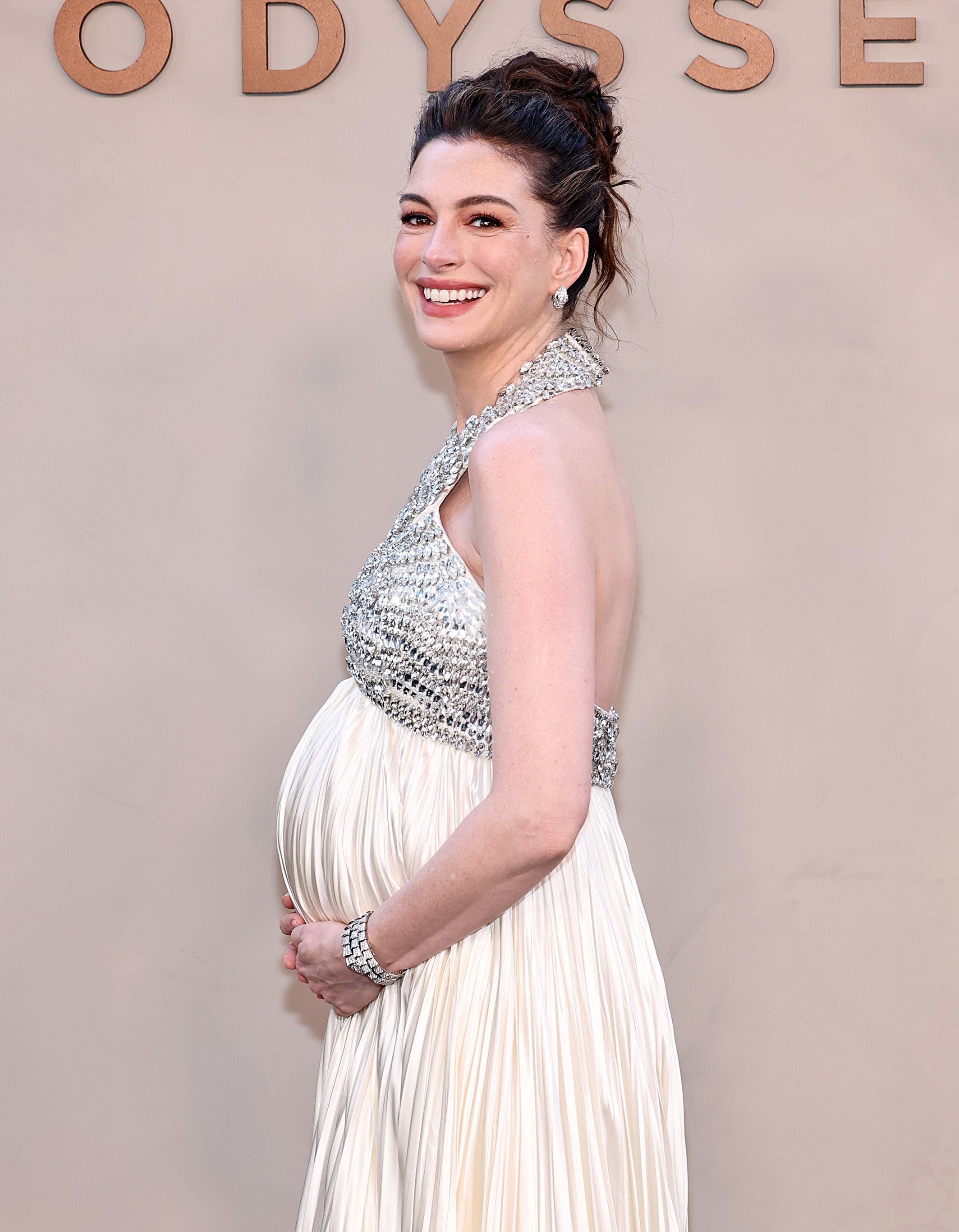 Person in elegant halter gown, embellished with sparkling details, poses cradling a visible baby bump, smiling on a Celebrity event background