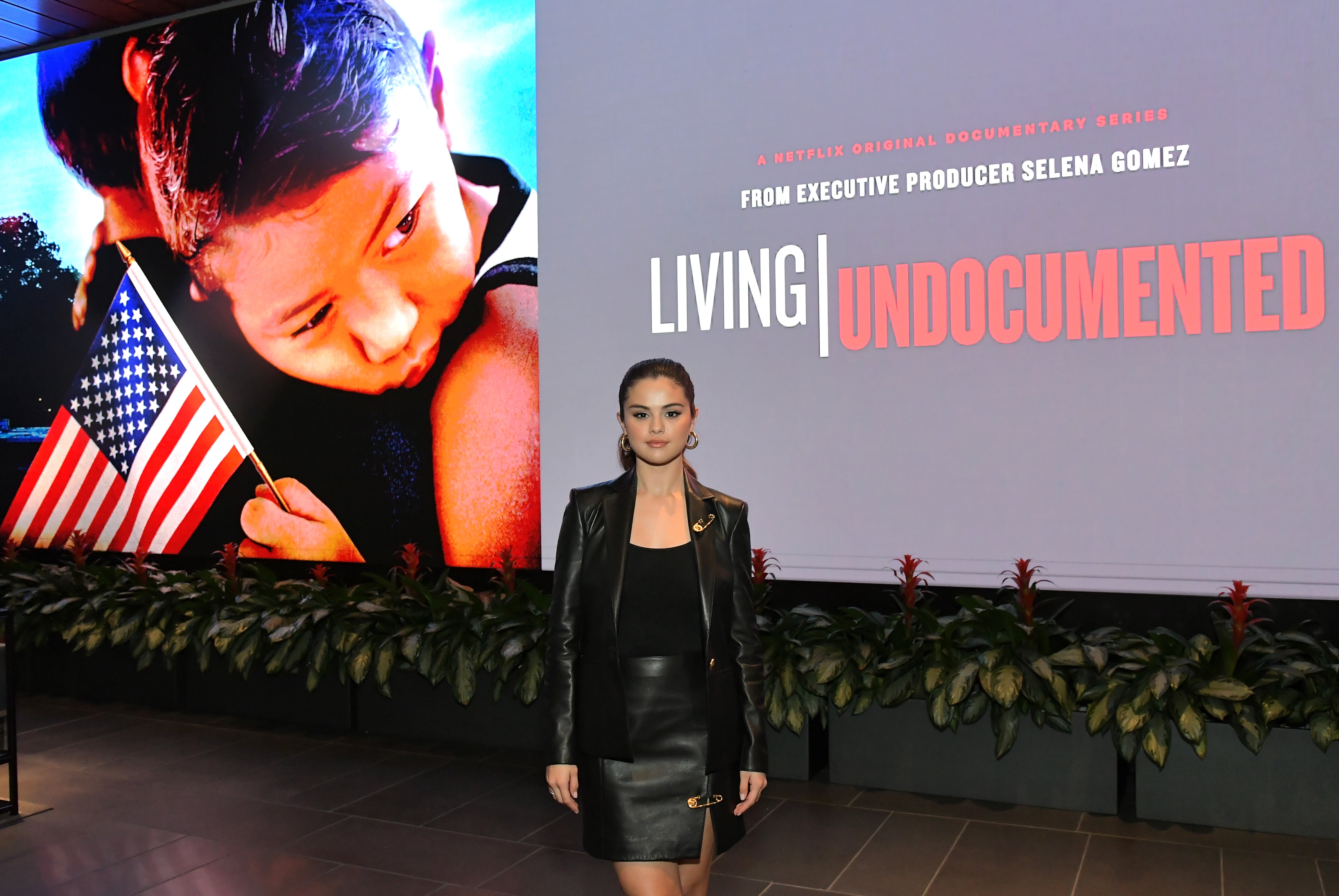 Selena Gomez in a leather ensemble stands before the "Living Undocumented" documentary backdrop.