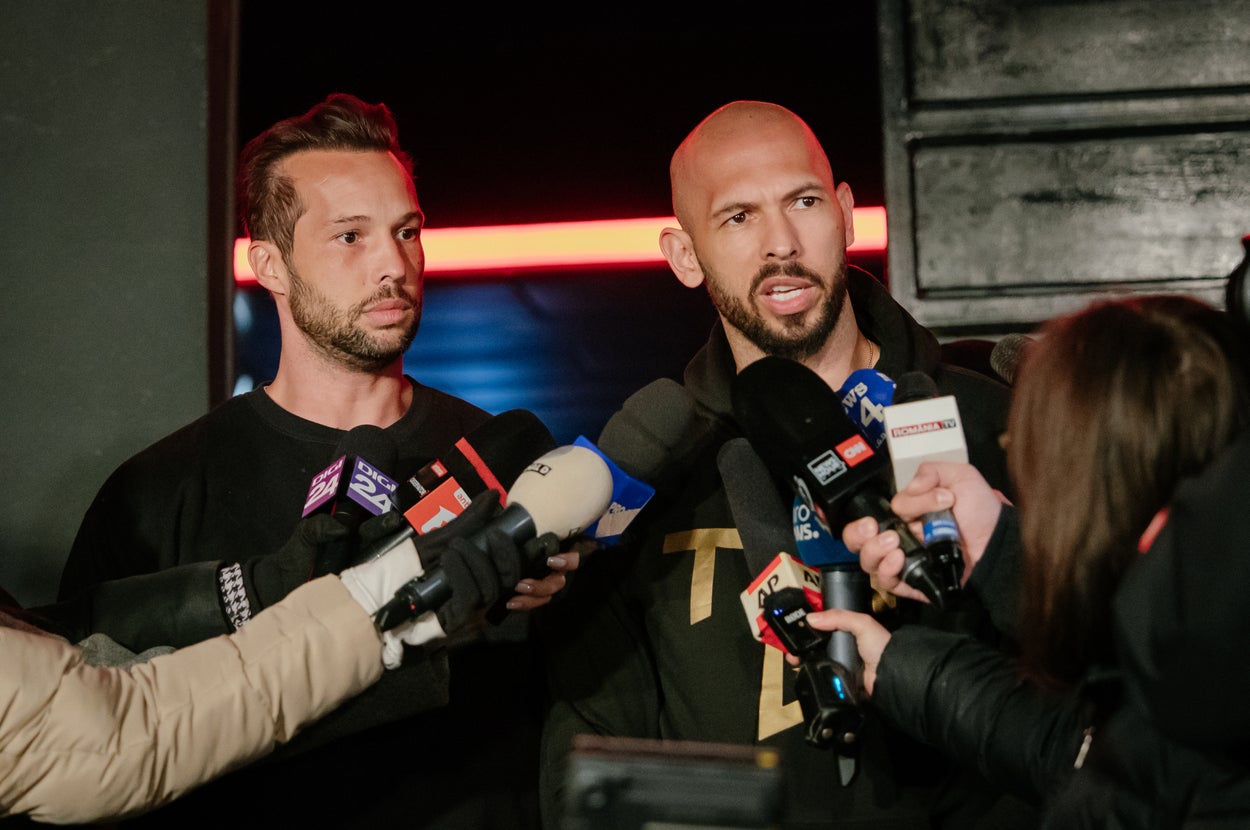 Two men surrounded by reporters holding microphones during a media interview