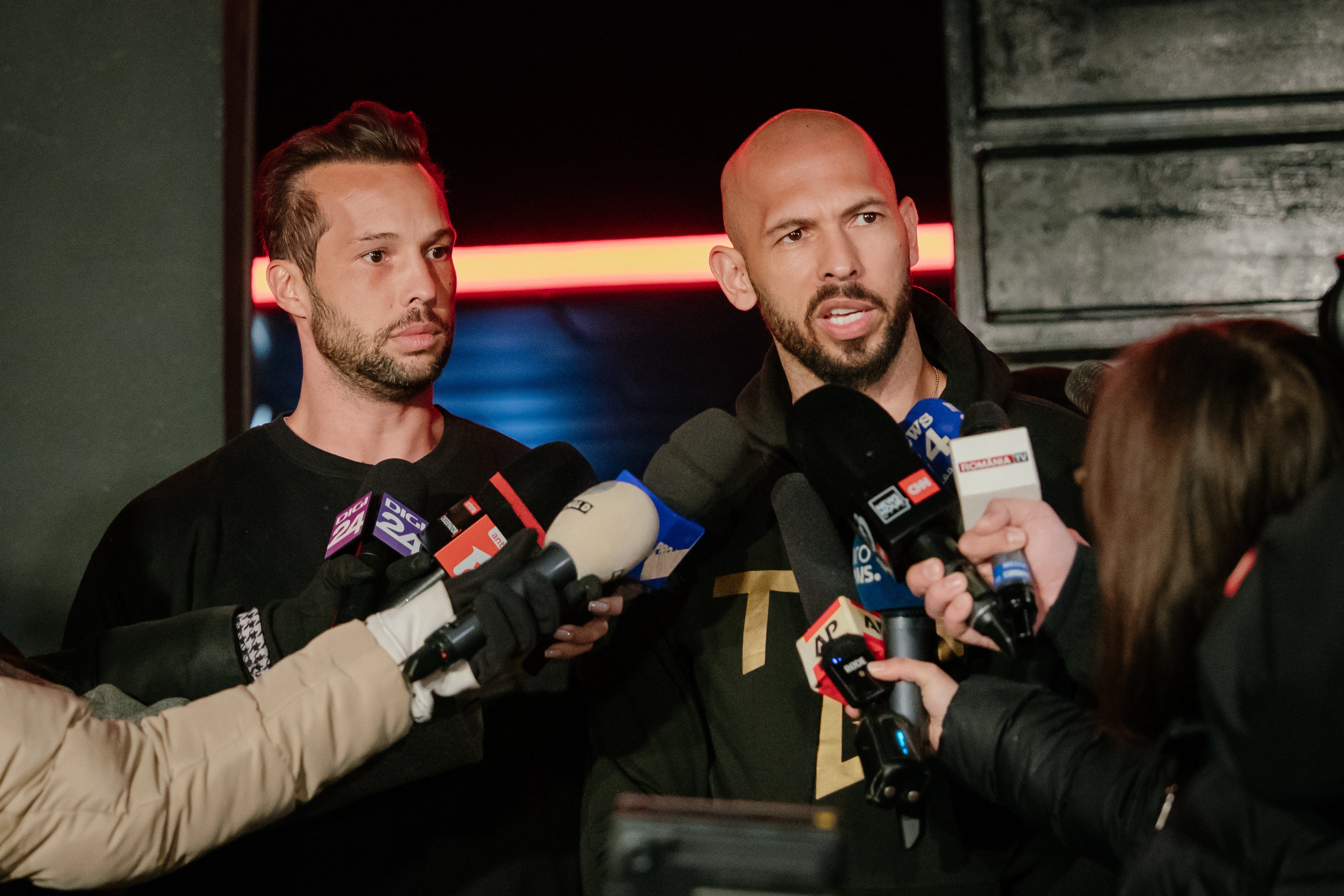 To menn holder et medieintervju, omgitt av mikrofoner. De har på seg uformelle mørke klær og engasjerer seg med journalister som stiller spørsmål