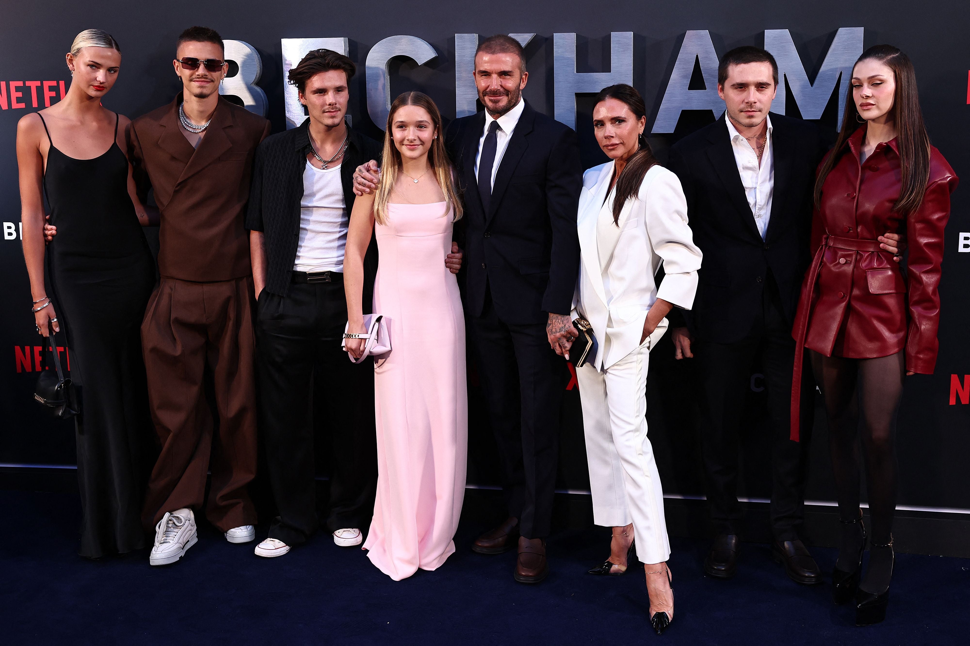 Group of eight people posing on a Netflix event red carpet. They're stylishly dressed: suits, a pink gown, white outfit, and a leather jacket