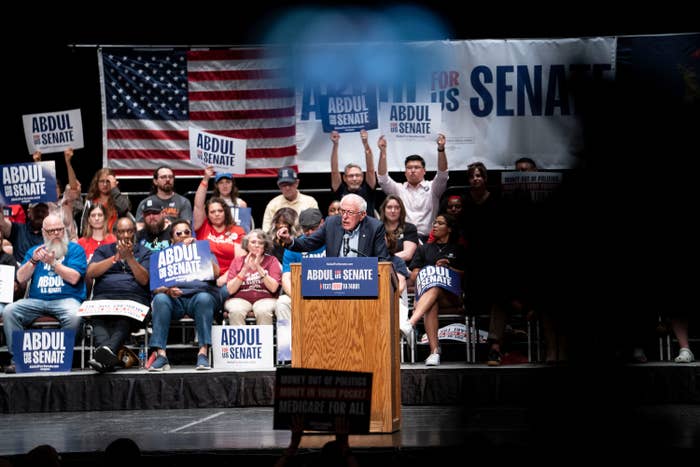 A speaker at a podium addresses a crowd at a political rally. Attendees hold signs reading "Abdul for U.S. Senate" with a U.S. flag in the background