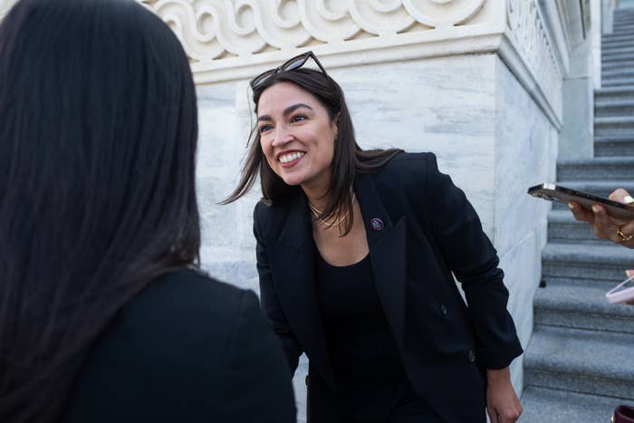 A person in a black outfit and glasses on head smiles engagingly at another person near marble steps, while someone nearby holds a phone