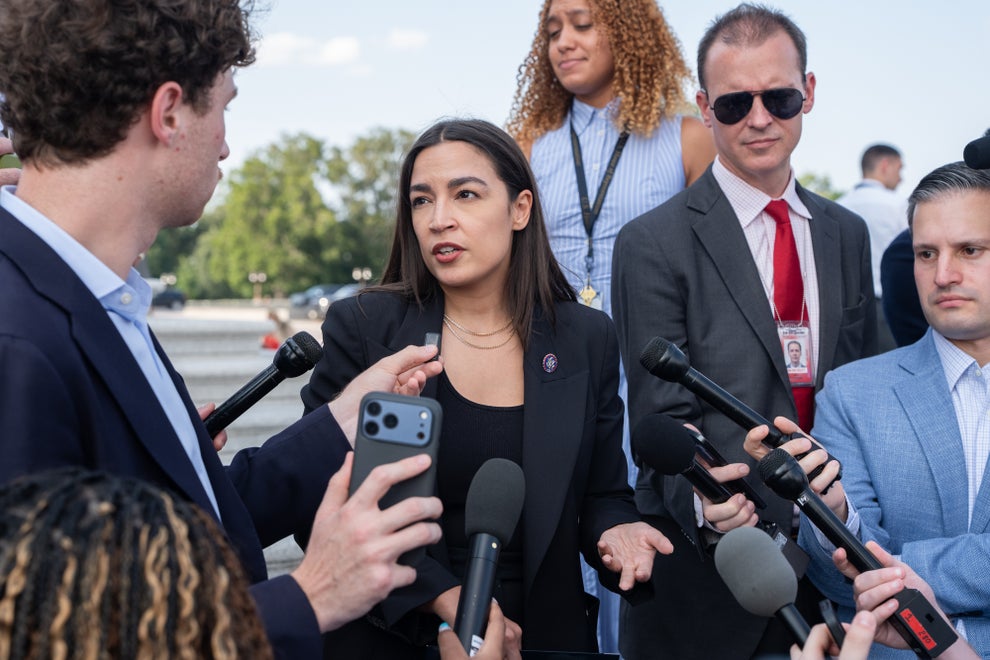 Politician speaking to reporters and surrounded by microphones, wearing a formal blazer, with spectators and media personnel in the background