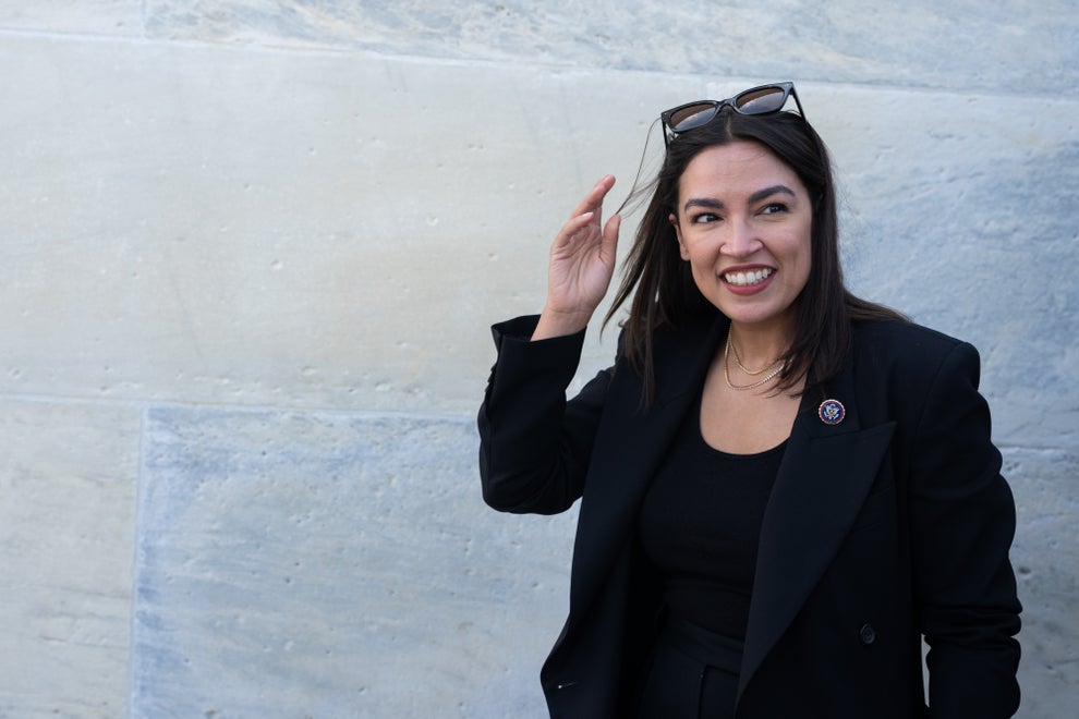 Person smiling and adjusting sunglasses outdoors, wearing a black blazer over a black top, standing against a stone wall