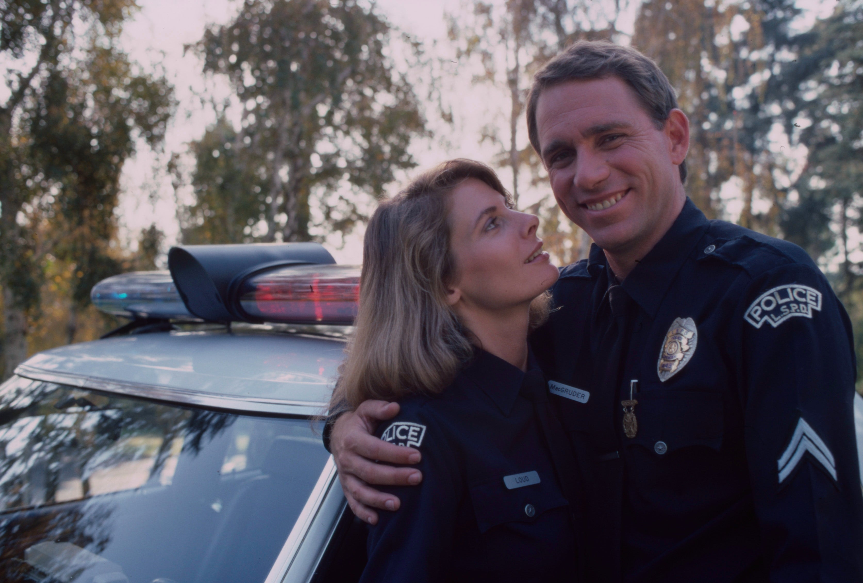 Two actors in police uniforms stand beside a patrol car, smiling. They appear to be portraying characters in a TV or movie scene
