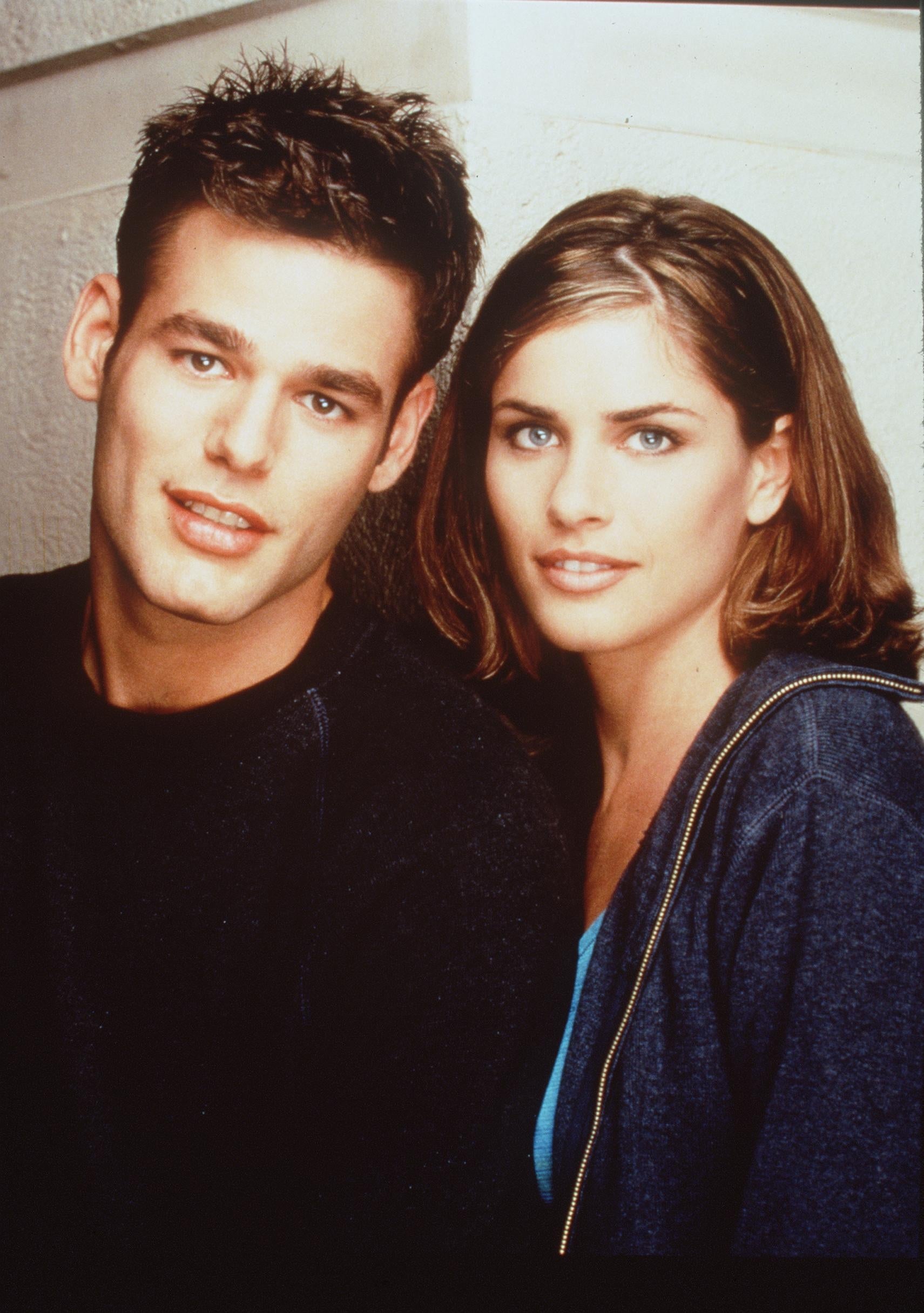 Two people pose closely together. The person on the left wears a dark top, and the person on the right wears a dark jacket over a blue shirt