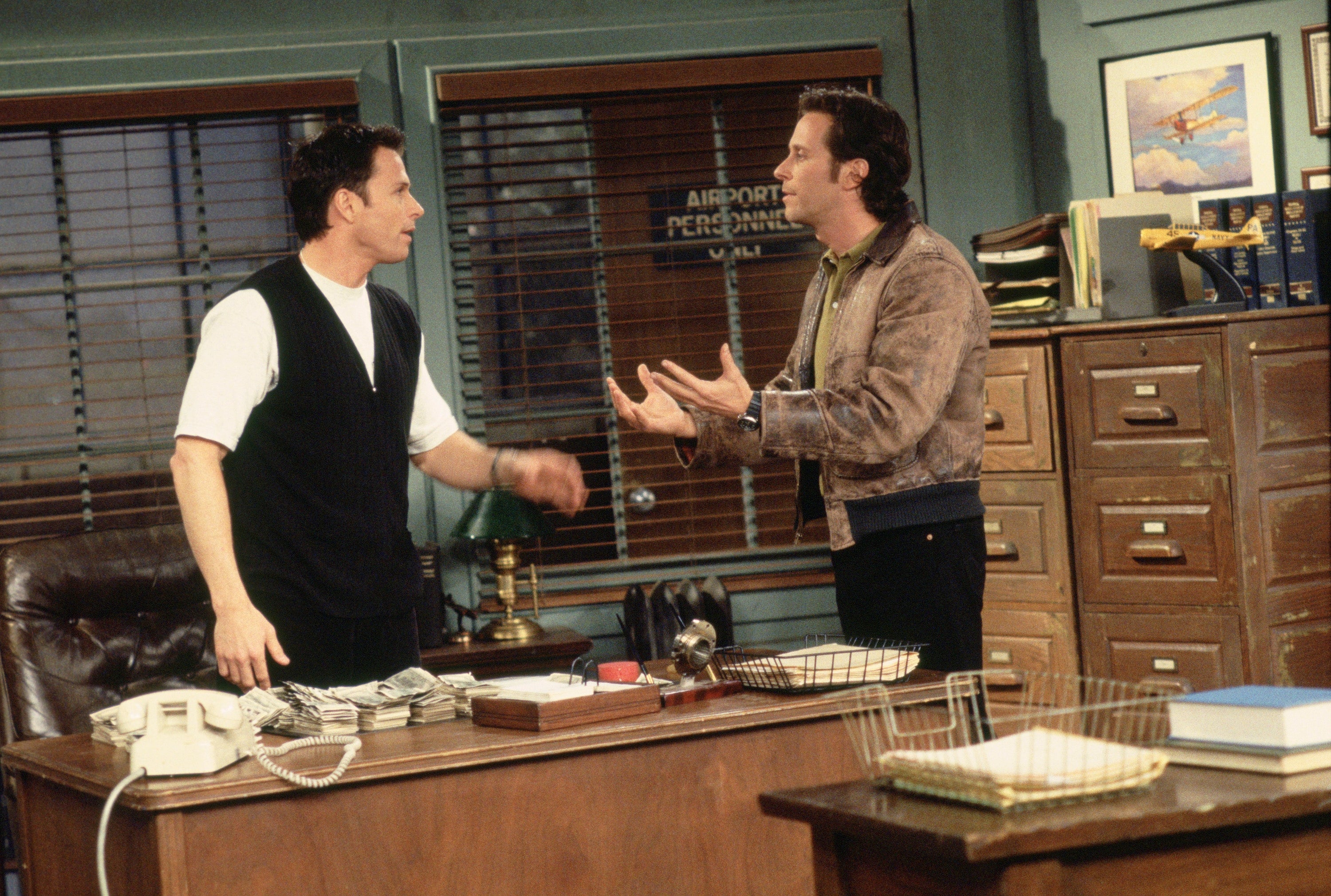 Two men in a TV show scene argue in an office with desks, shelves, and papers around them. One man wears a jacket, the other a vest