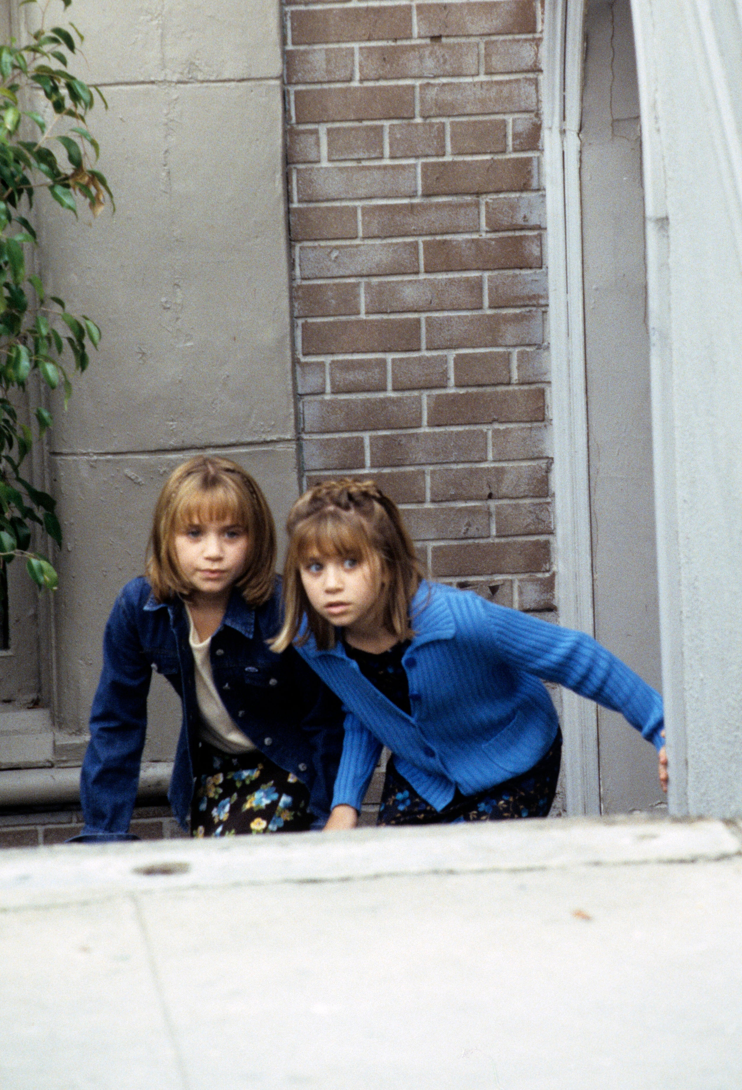 Two young girls appearing curious, peering around a corner outside a building