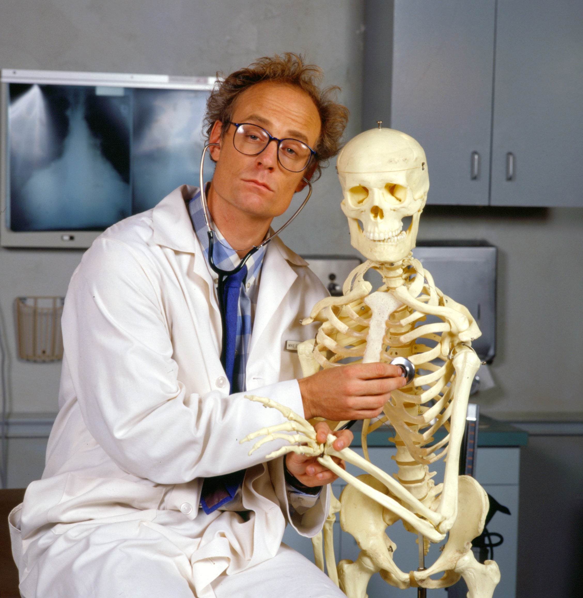 A person in a lab coat humorously uses a stethoscope on a skeleton, suggesting a playful take on a medical scene