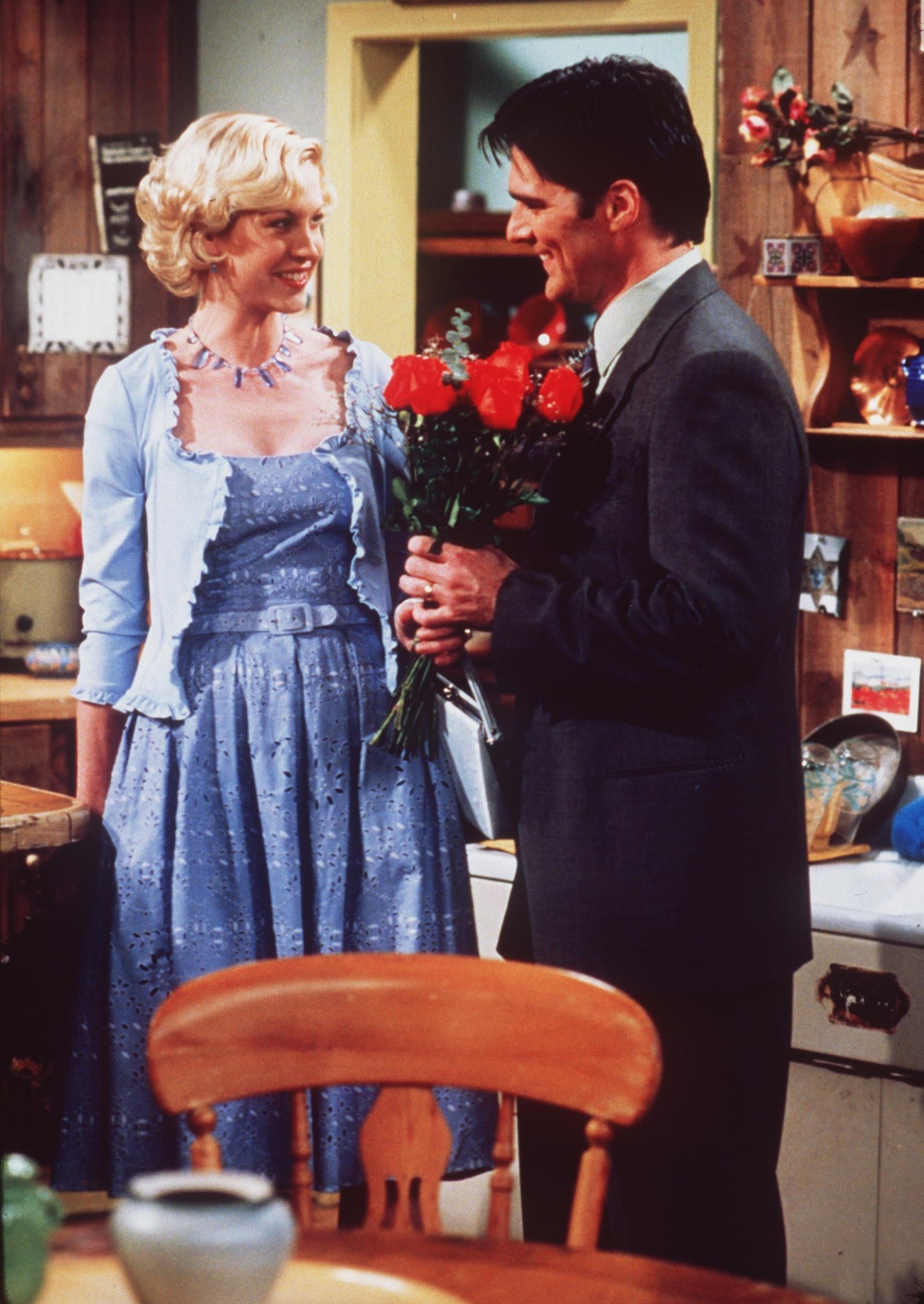 Two actors in a sitcom scene: a woman in a patterned dress with a cardigan smiles at a man in a suit holding a bouquet of roses
