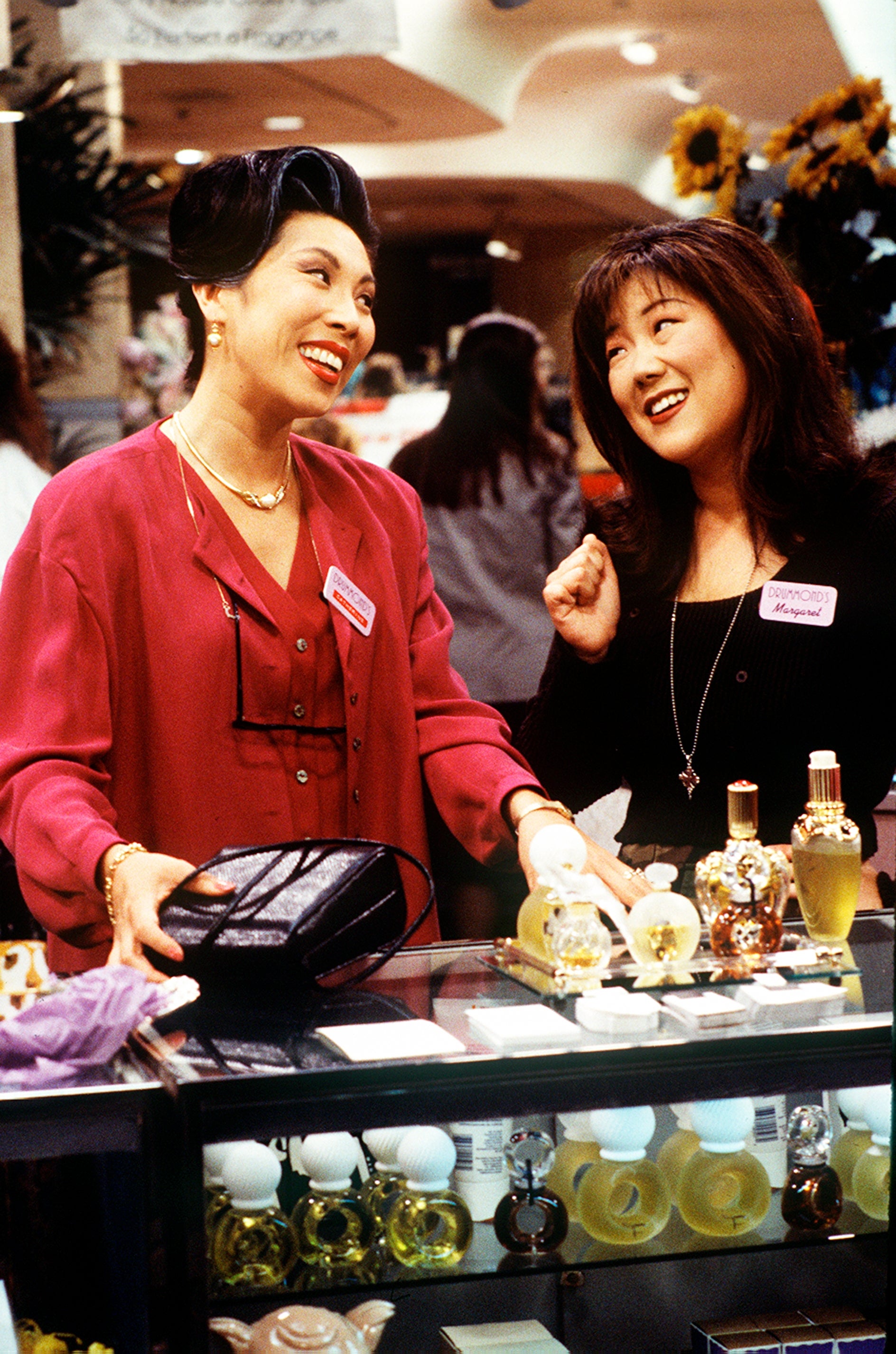 Two women, smiling and interacting at a store counter, standing near perfume bottles, both wearing name tags and casual attire