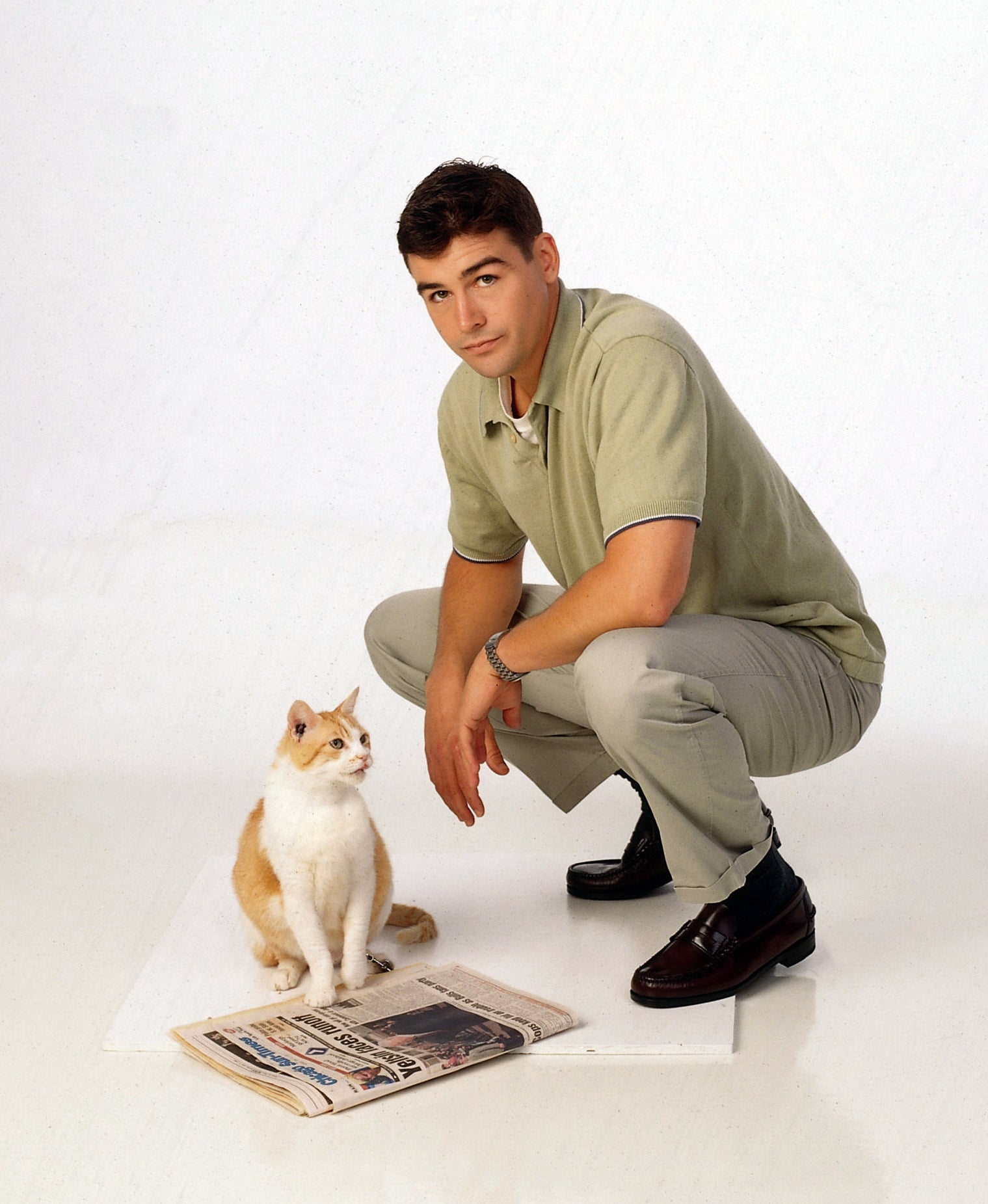 Person squatting next to a cat sitting by a newspaper on the floor, both facing forward