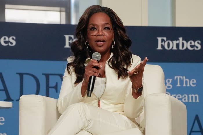 A woman in a stylish white suit speaks on a panel, holding a microphone, with a "Forbes" backdrop