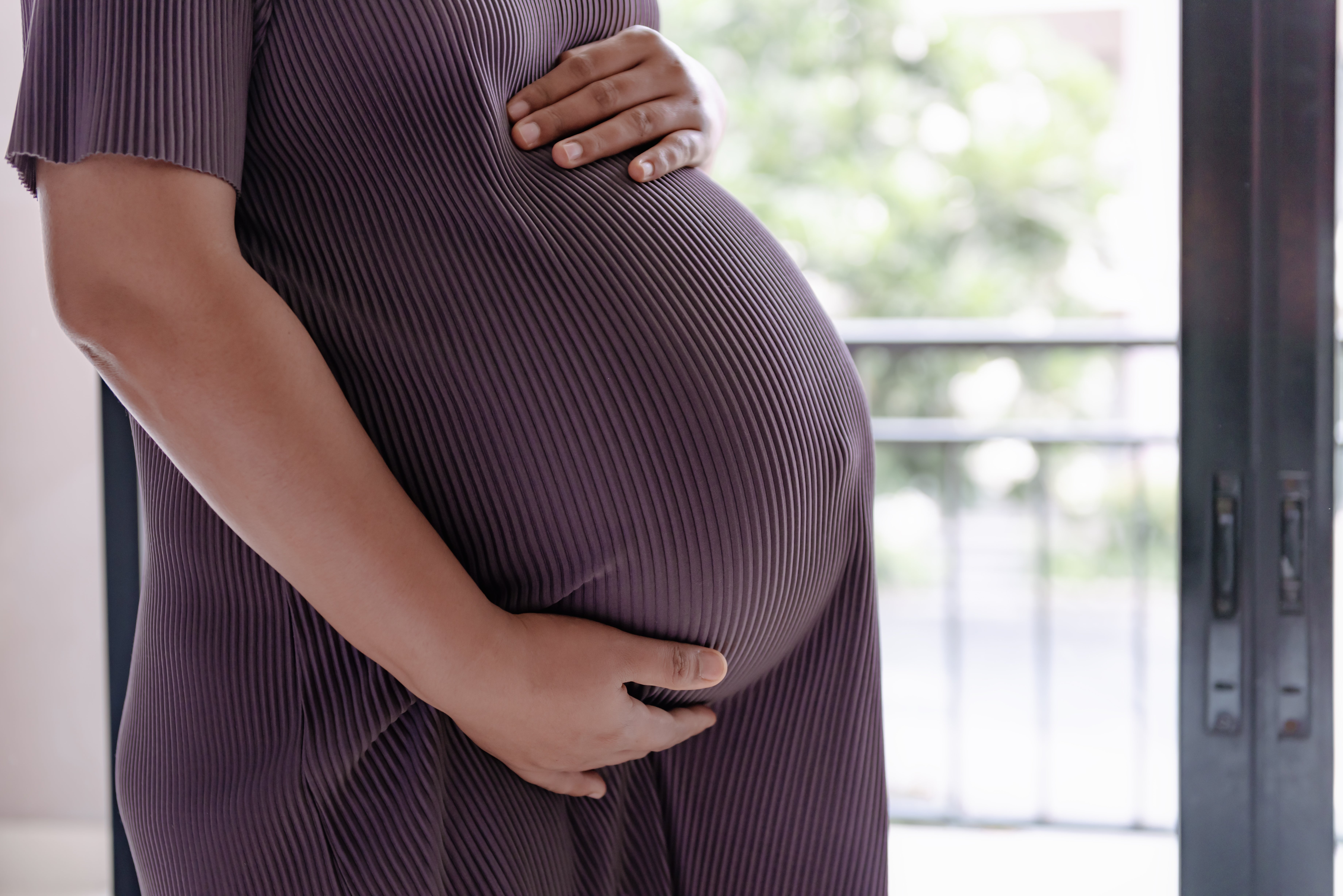 Gravid person i ribbet kjole holder forsiktig magen og står nær et vindu