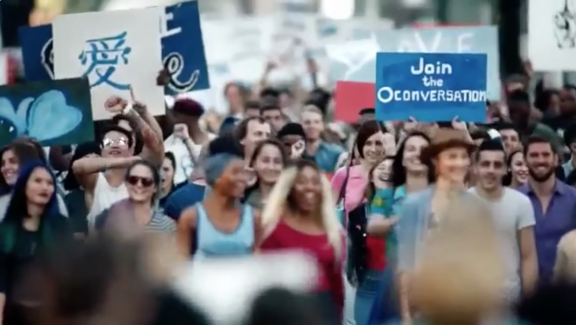 A crowd participates in a public demonstration, holding signs with various messages, including "Join the Conversation."
