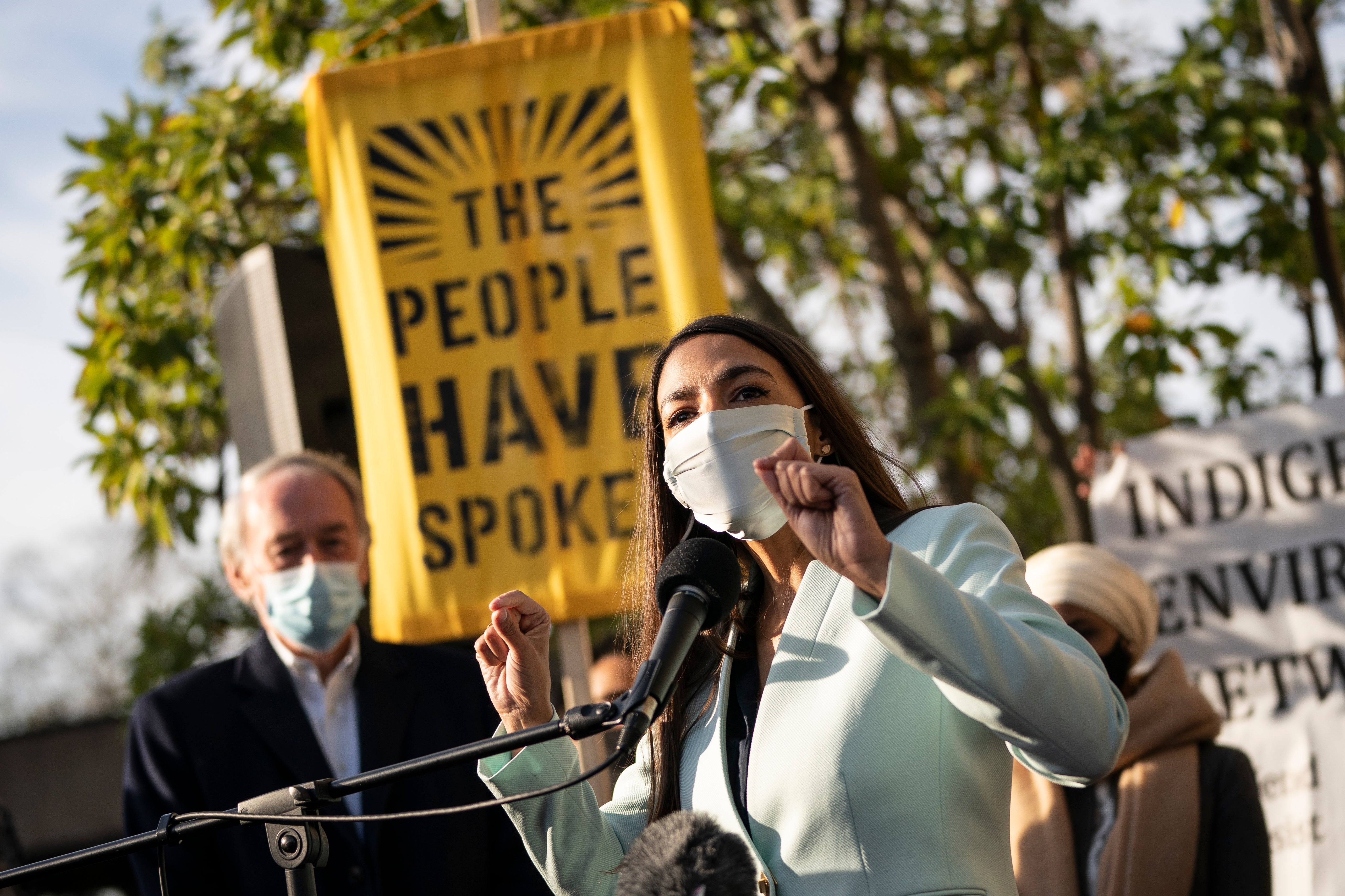 Alexandria Ocasio-Cortez speaking outside of the Democratic National Committee headquarters on a podium. A banner behind reads, "The People Have Spoken."
