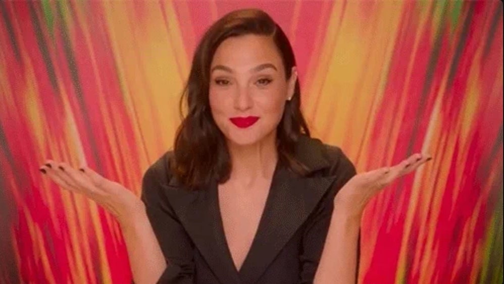 Gal Gadot smiles while throwing her hands up, sitting against a vibrant backdrop.