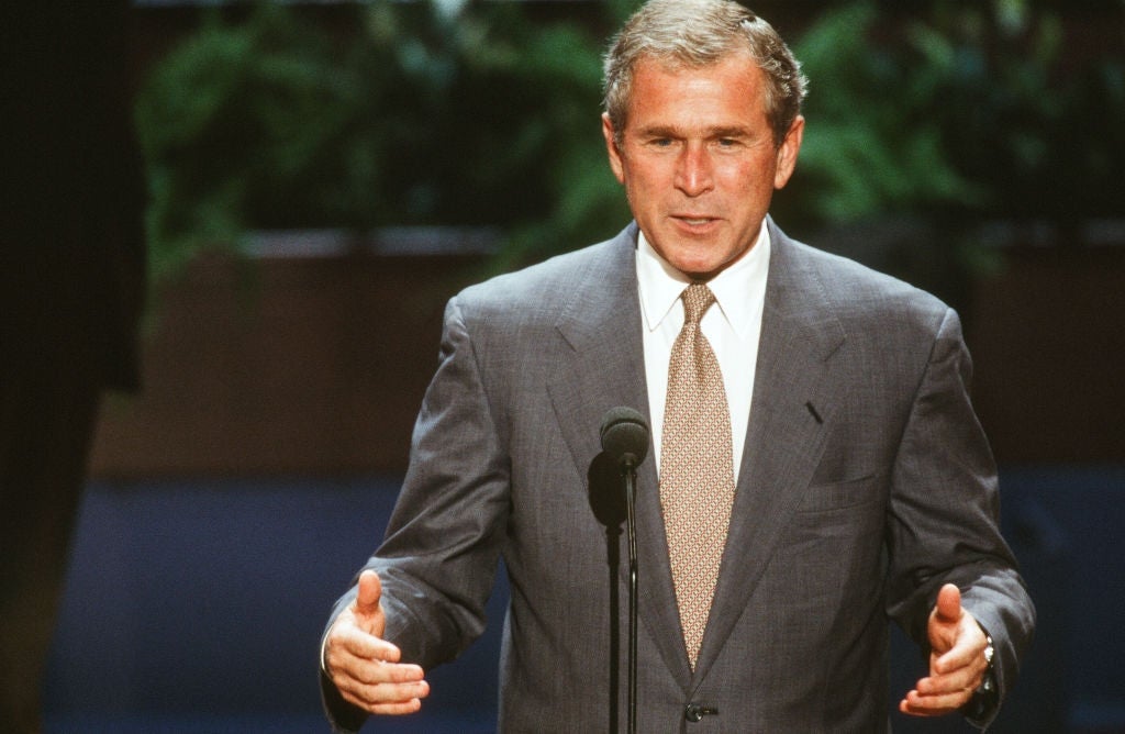 George W. Bush i dress taler ved et podium, gestikulerer med hendene, i en formell setting