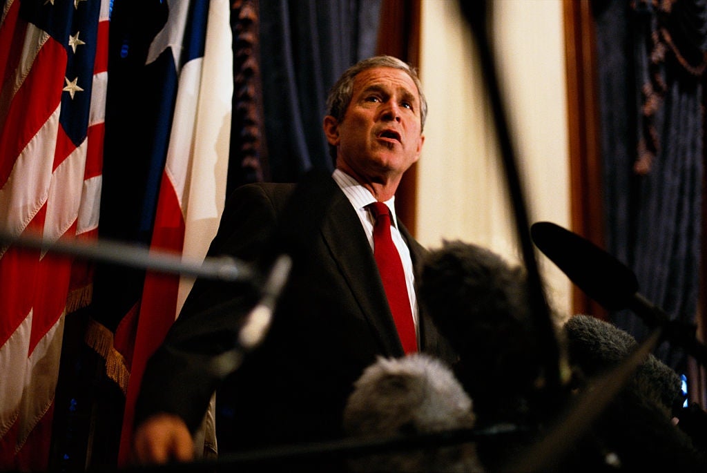 George W. Bush i dress snakker inn i flere mikrofoner på en konferanse, med flagg i bakgrunnen