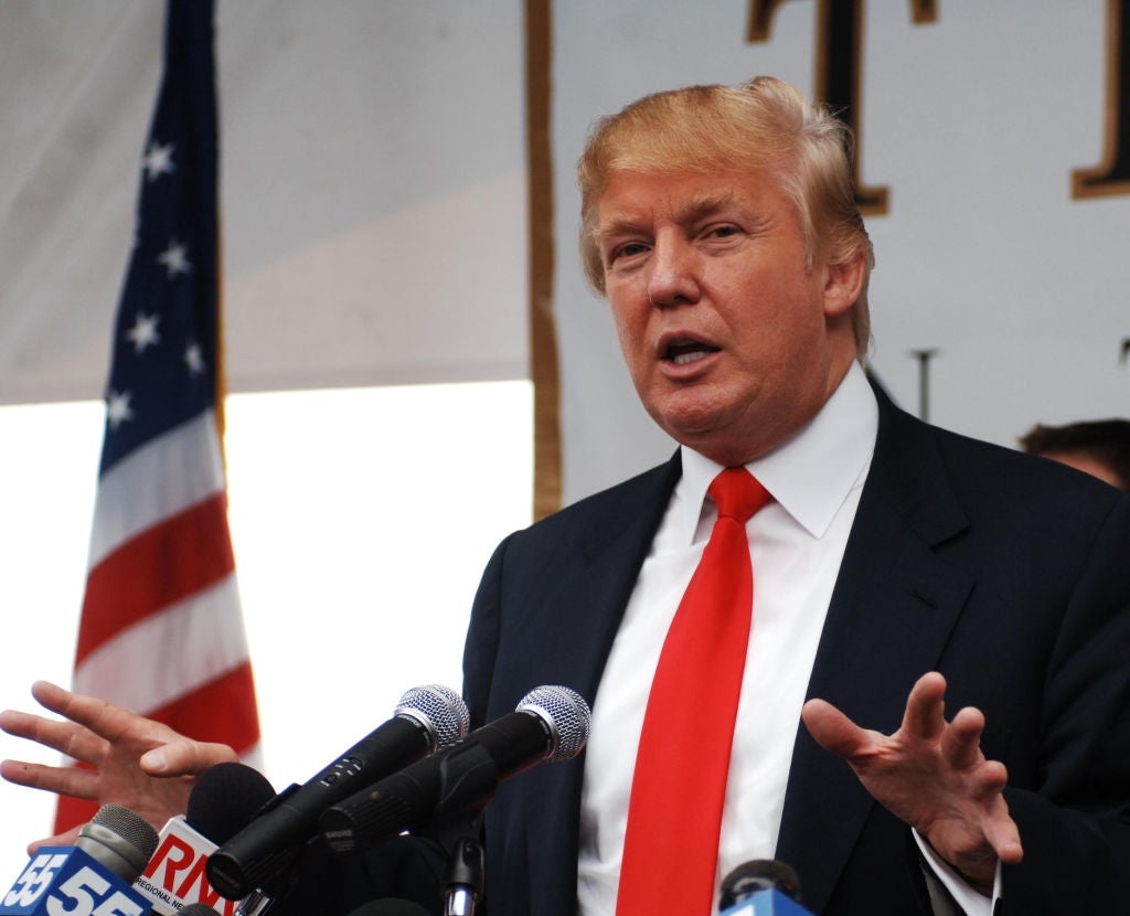 Donald Trump i dress med slips snakker på en pressekonferanse, stående bak flere mikrofoner