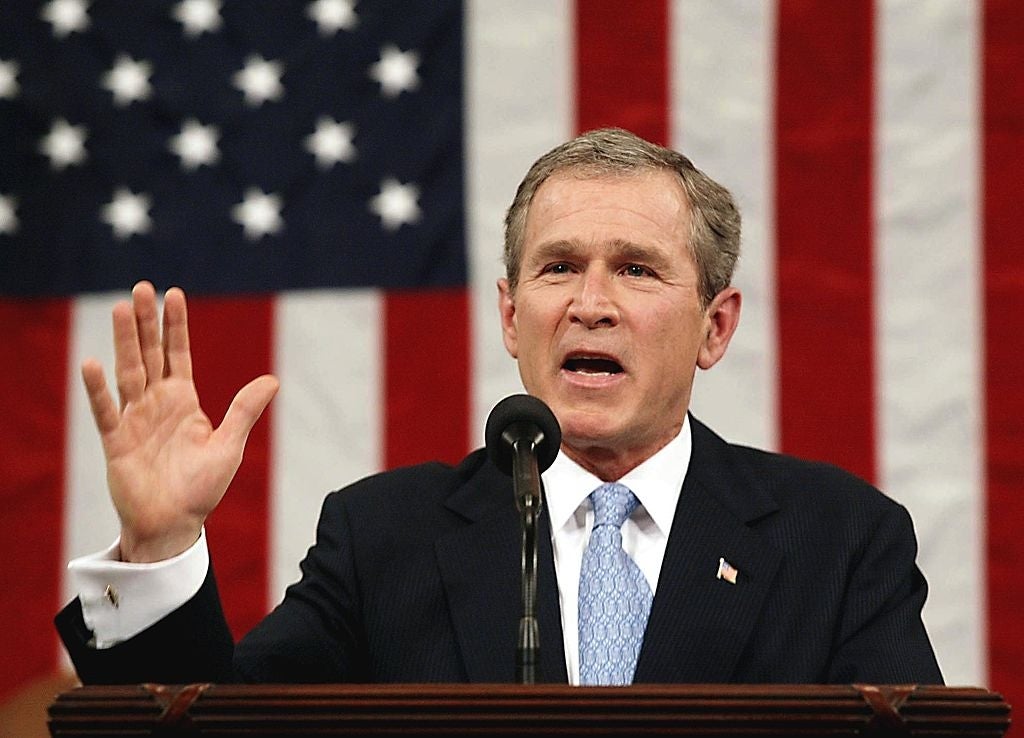 George W. Bush, i dress, taler ved et podium med et amerikansk flagg i bakgrunnen. Han har en hånd løftet mens han henvender seg til et publikum
