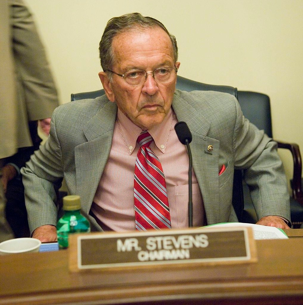 Ted Stevens i dress og stripet slips sittende ved et bord med navneskilt