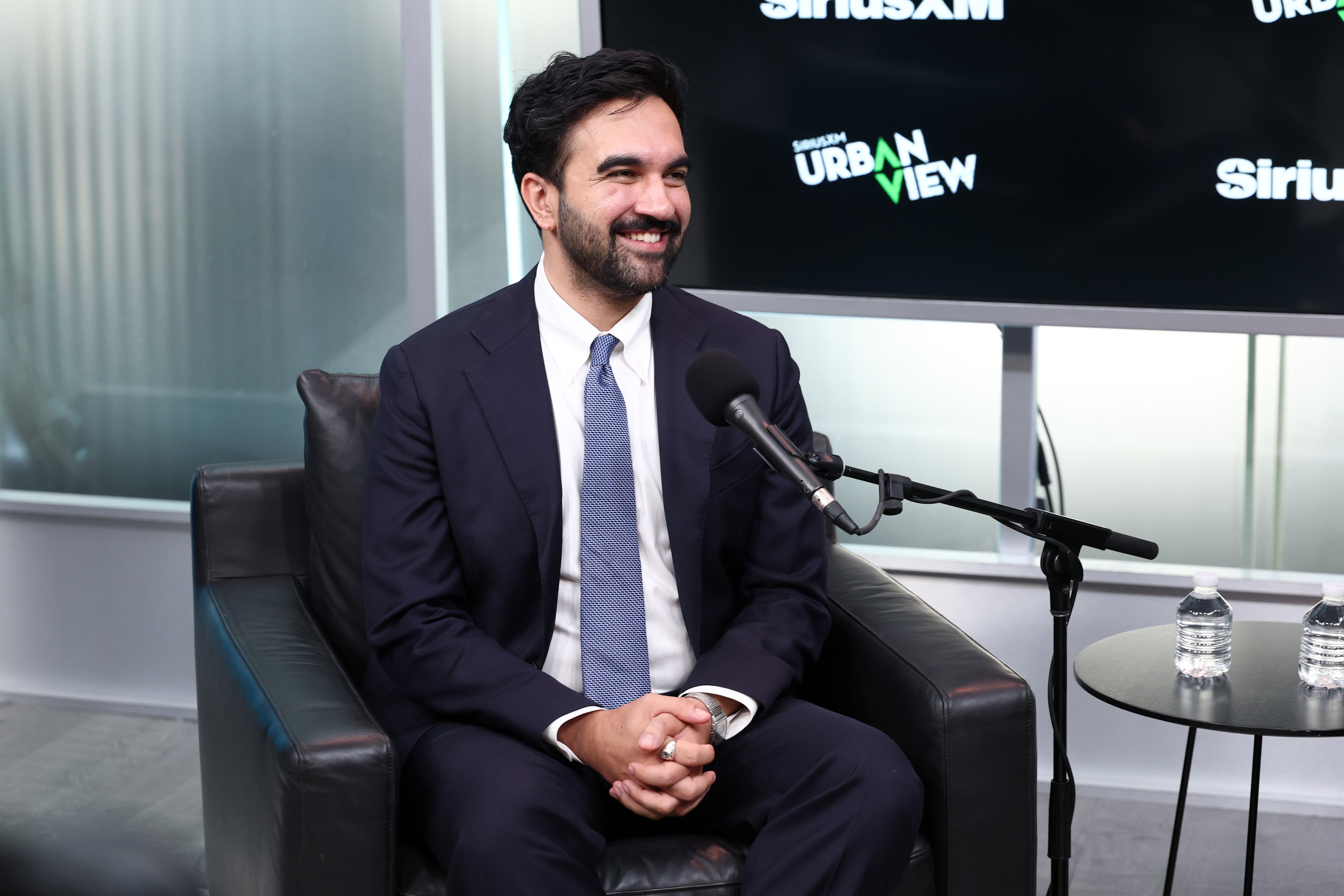 En person i dress og slips sitter i et studio, smilende, med en mikrofon foran. Vegger og skjerm har "SiriusXM" og "Urban View" logoer